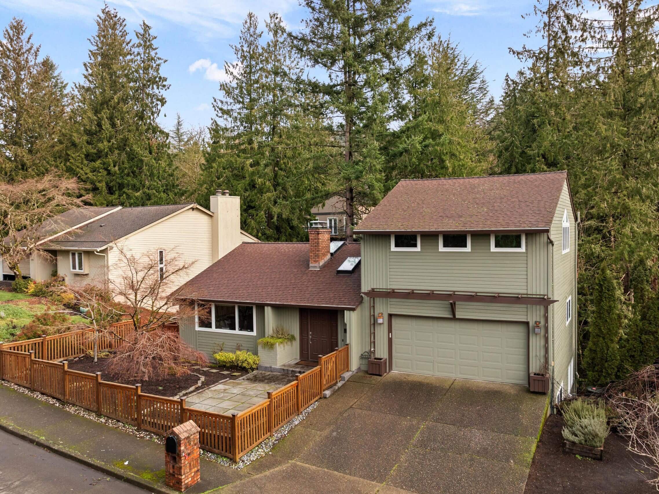 A two-story suburban house with green siding, a brown roof, attached garage, fenced front yard, and patio, surrounded by tall evergreen trees and neighboring homes. The driveway and street are visible in the foreground.