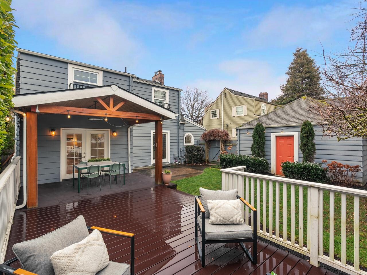 A backyard with a wooden deck featuring outdoor seating, a covered dining area, a green lawn, and two blue-gray houses with white trim. There is also a small shed with a red door and surrounding landscaping.