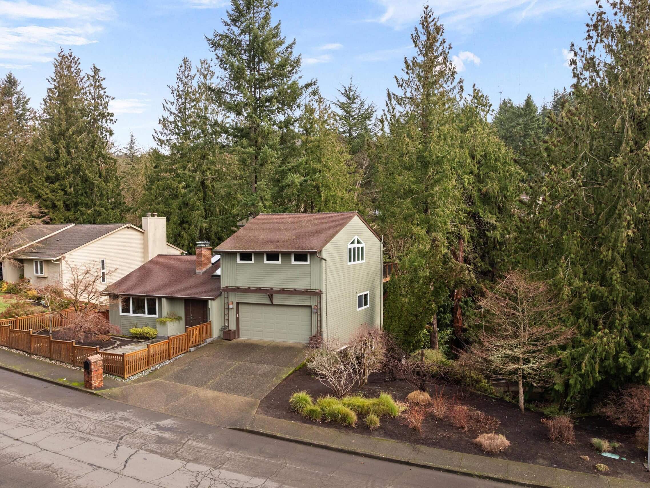 Two-story house with a brown roof and attached garage, surrounded by tall evergreen trees and landscaped shrubs, located on a quiet suburban street.