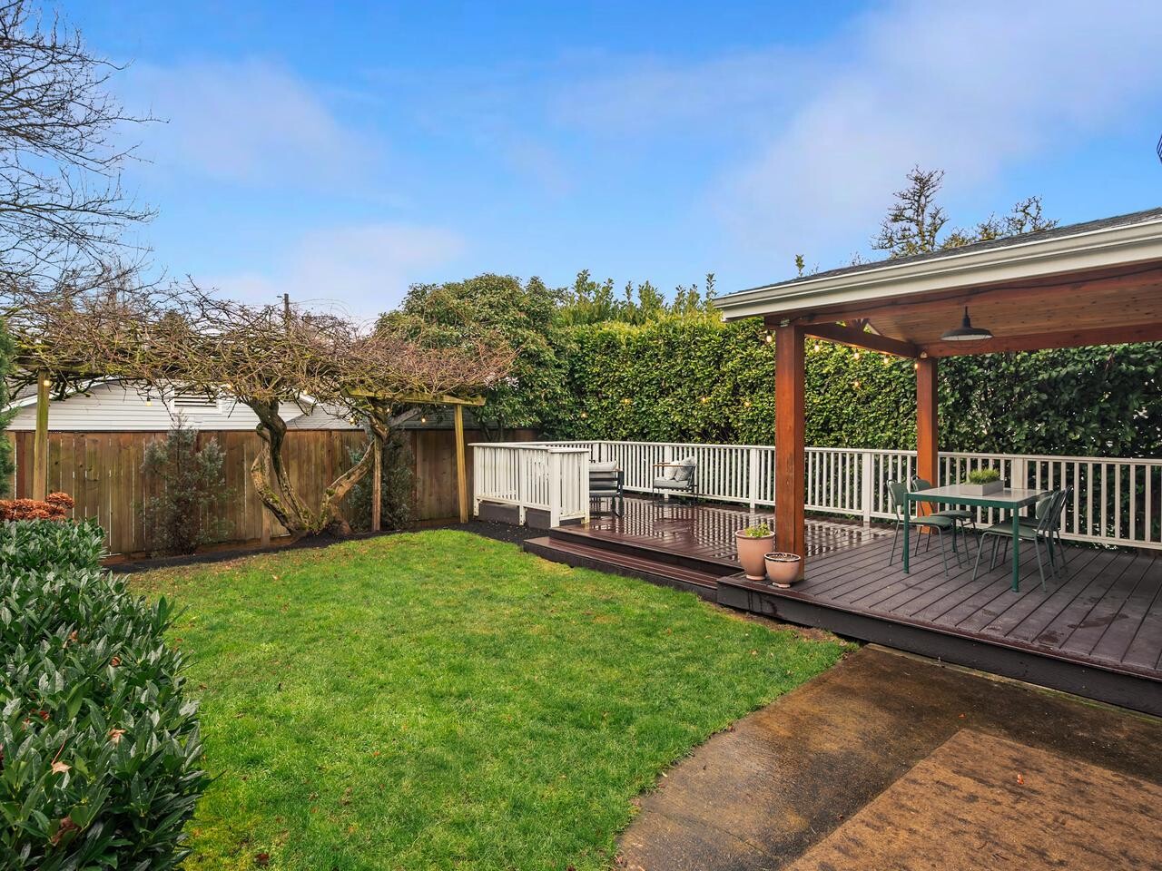 A backyard with a covered wooden deck featuring outdoor furniture, a green lawn, shrubs, and a tree along a wooden fence under a partly cloudy blue sky.