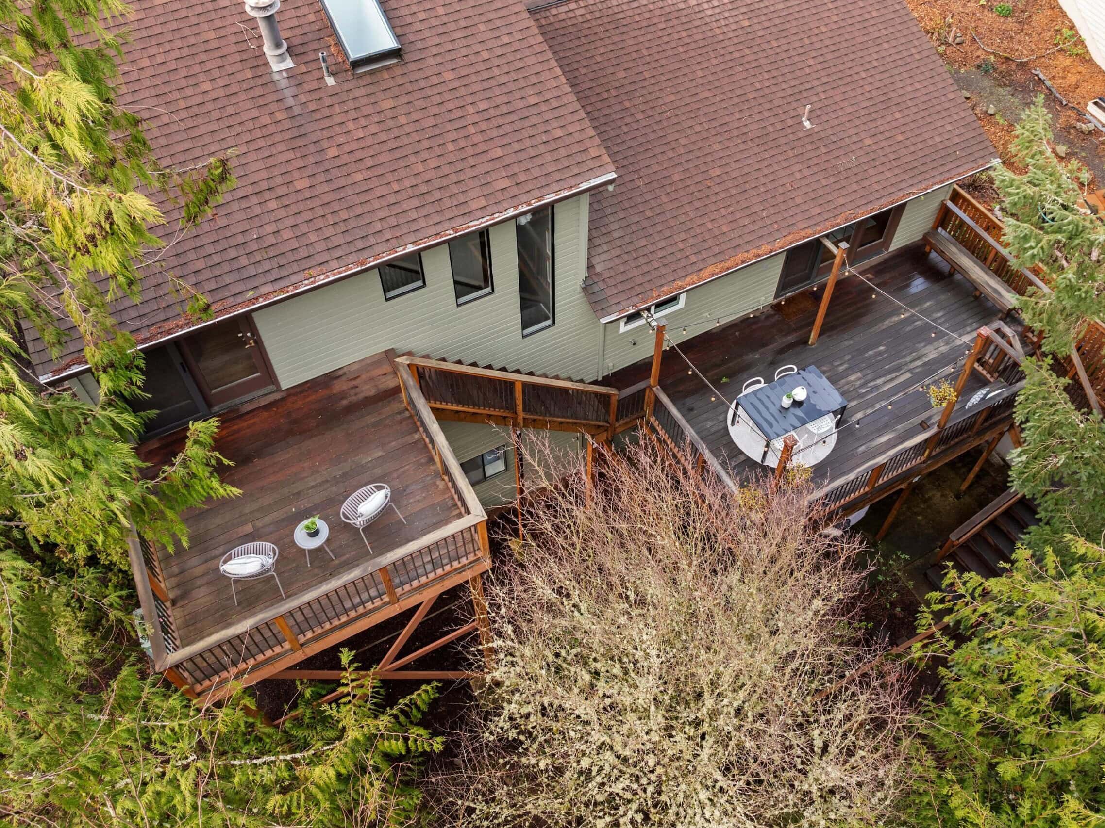 Aerial view of a house with two wooden decks surrounded by tall trees. One deck has two chairs and a small table, while the other features a round table with chairs. The decks and house are nestled in a lush, forested area.
