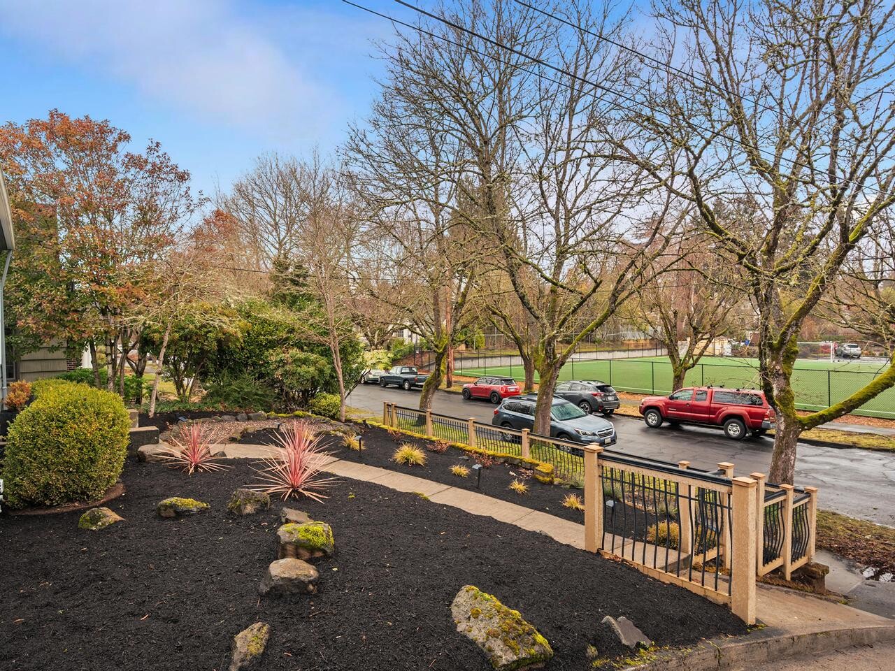 A sloped front yard with dark mulch, scattered rocks, and small plants, bordered by a wooden fence, next to a sidewalk and street with parked cars and leafless trees in front of a sports field.