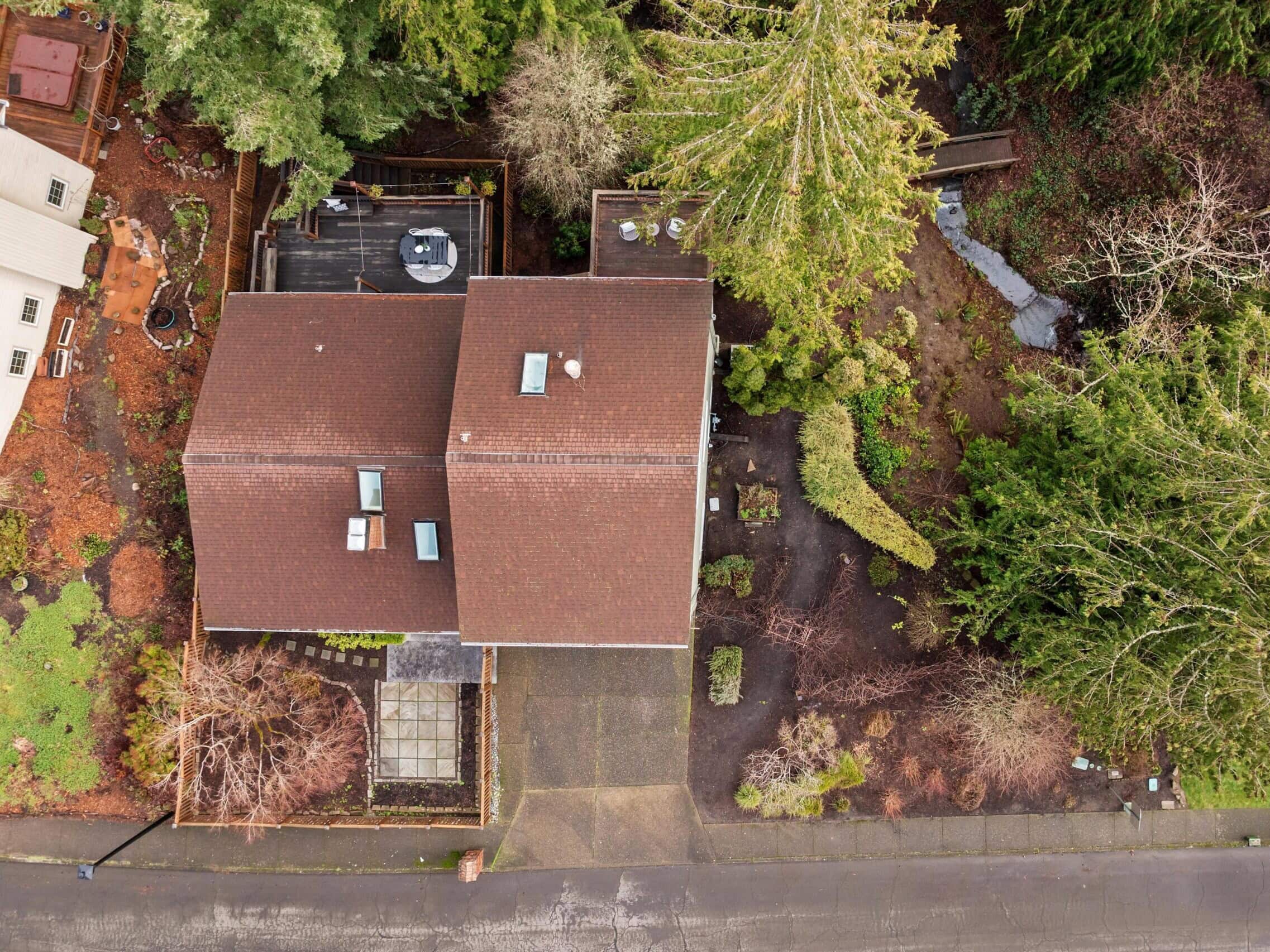 Aerial view of a house with a brown roof surrounded by trees and gardens, adjacent to a road. The yard includes a patio, walkways, and various shrubs and greenery. Neighboring house is partially visible on the left.