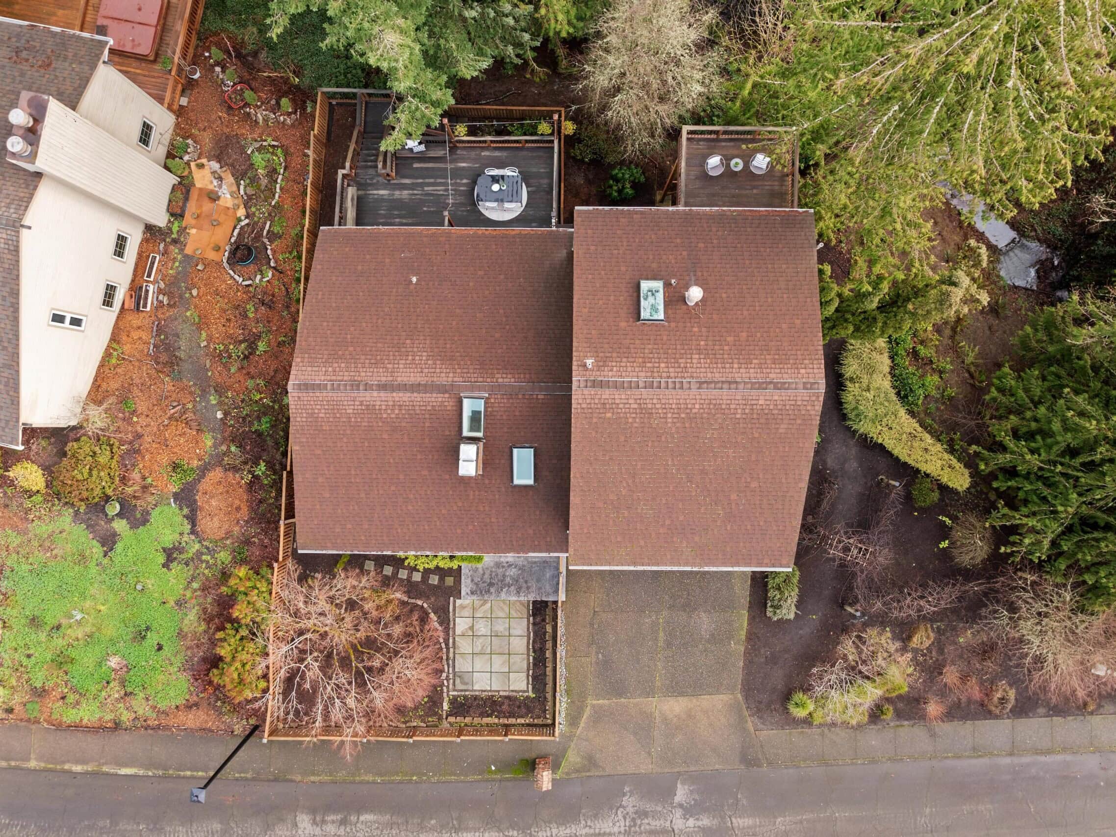 Aerial view of two houses with brown roofs surrounded by trees and gardens. One house has a deck with outdoor furniture, and the area includes a driveway, pathways, and green landscaping.