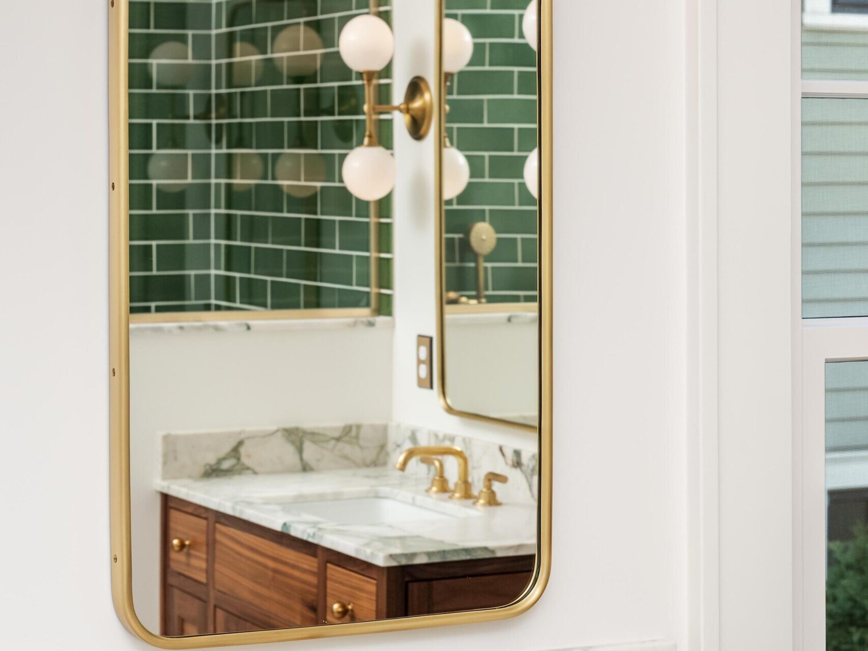 A bathroom vanity with a marble countertop, gold faucet, and a rectangular mirror. Above the mirror is a gold light fixture with two round bulbs. The reflection shows green subway tiles and a window.
