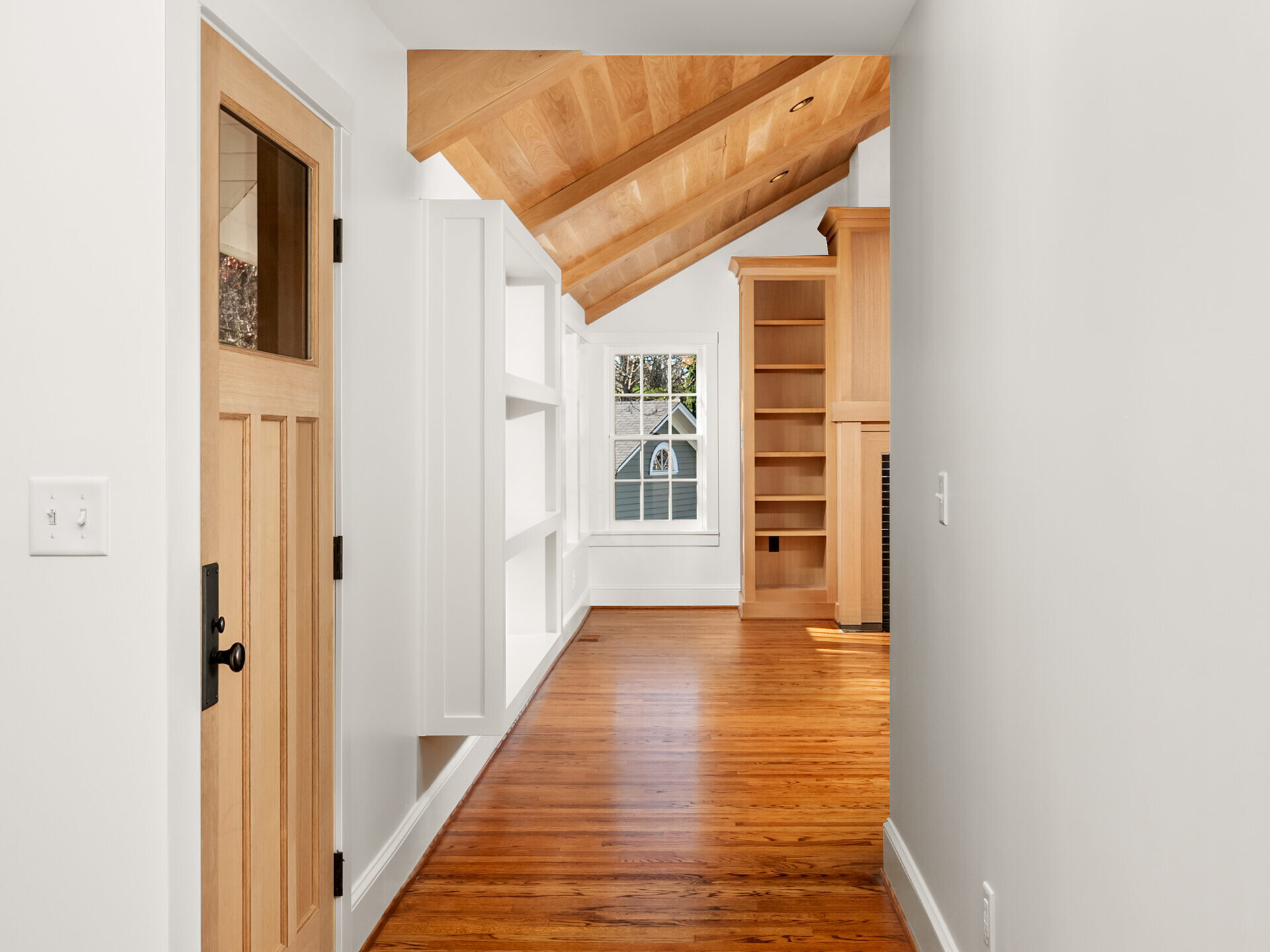 A bright hallway with wooden floors, white walls, built-in shelves, a wooden door on the left, and a vaulted wood-paneled ceiling leading to a sunlit room with large windows.
