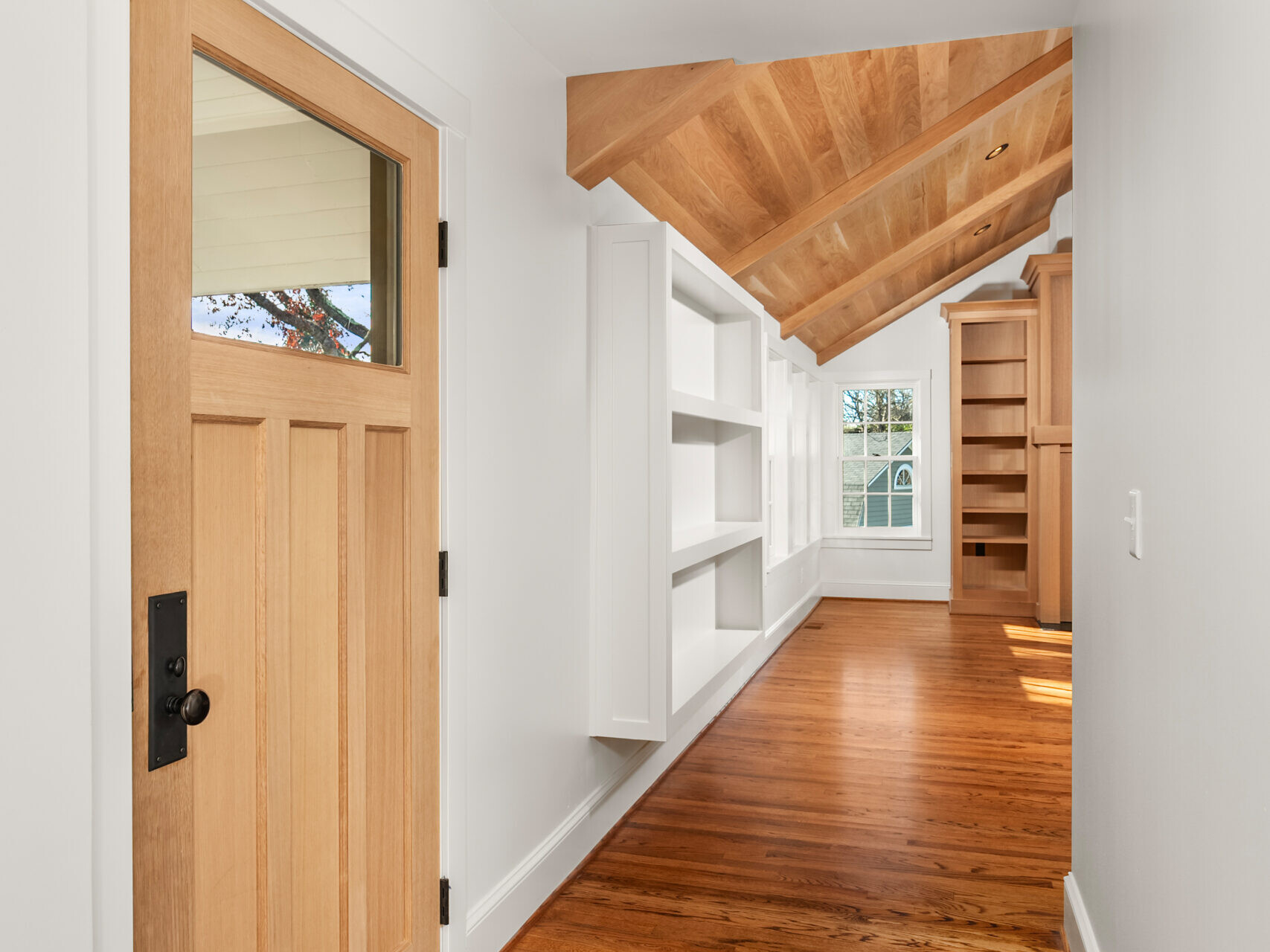 Bright hallway with wooden floors, a light wood door with a window, white walls, built-in white shelves, vaulted wood ceiling, and large window at the far end letting in natural light. A round ceiling light hangs above.