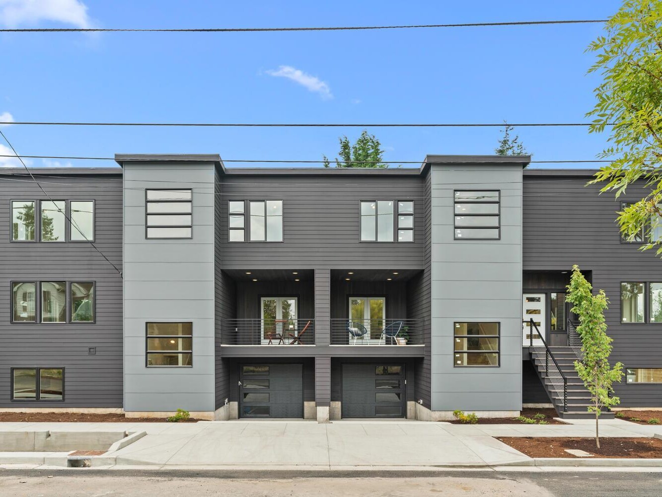 Modern gray multi-family townhouse with large windows, two stairways leading up to separate entrances, small balconies, and a clean, landscaped front area under a blue sky with a few clouds.