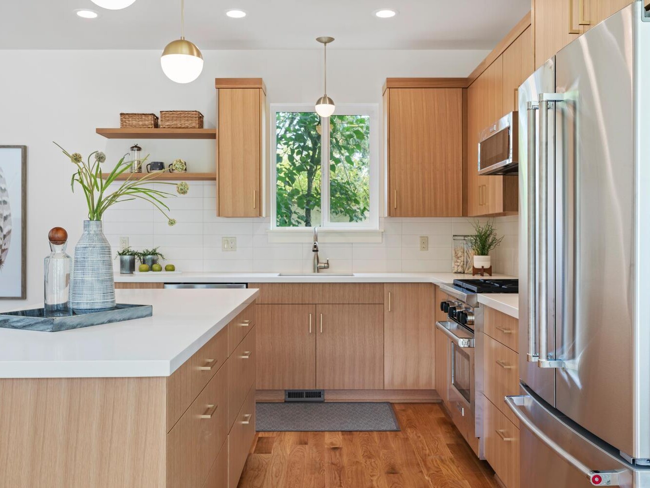 Modern kitchen with light wood cabinets, white countertops, stainless steel appliances, and a window above the sink. Minimalist decor includes vases, plants, and wall art with feathers. Natural light brightens the space.