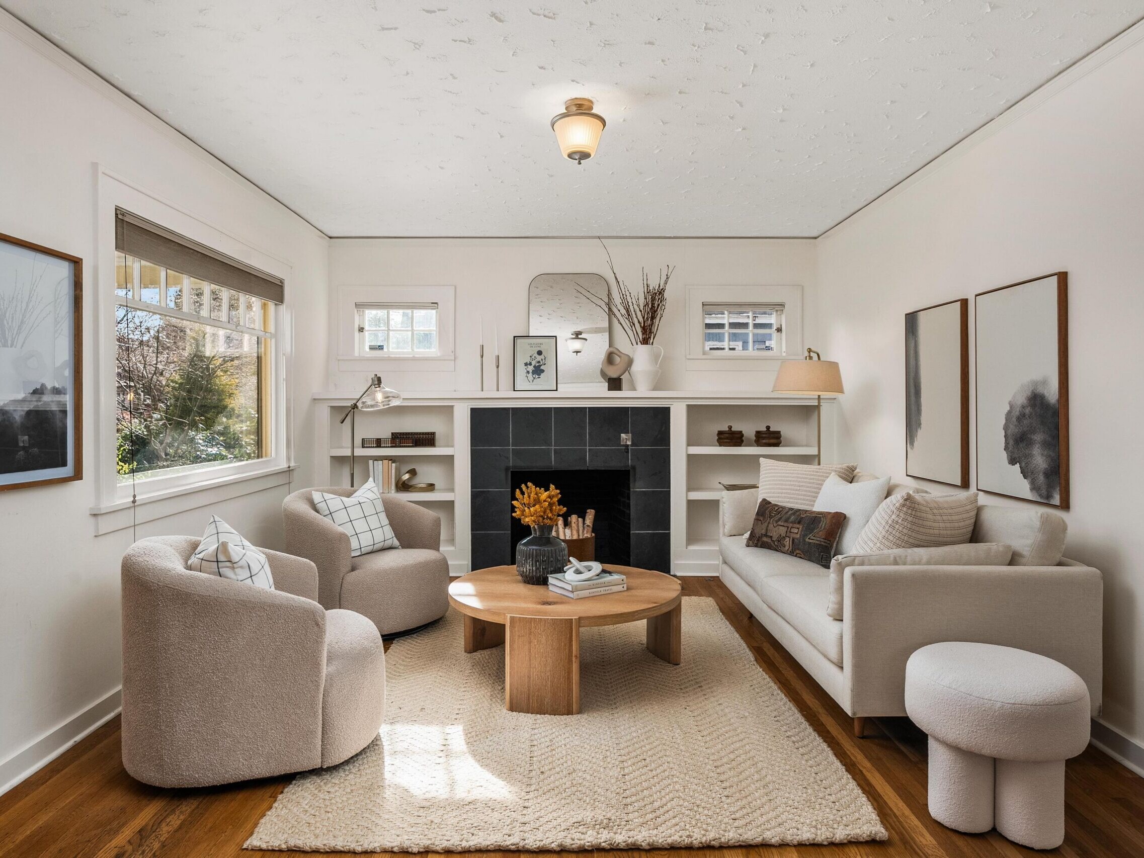 A cozy living room with neutral decor features a beige sofa, two round armchairs, a wooden coffee table, a fireplace with a black surround, built-in shelves, soft lighting, and minimalist wall art.