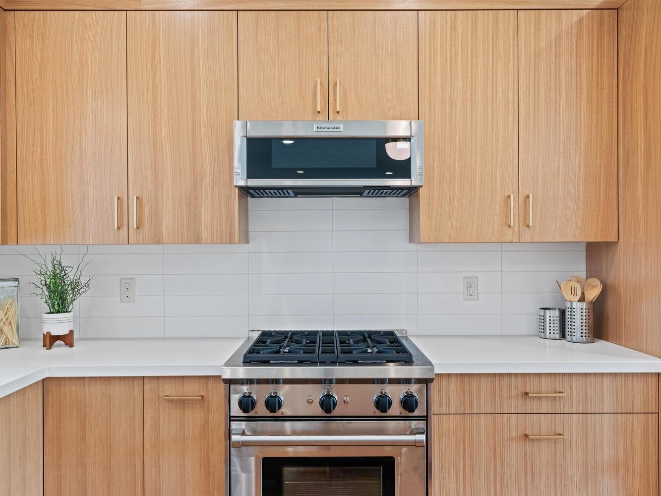 Modern kitchen with light wood cabinets, a stainless steel gas stove and oven, a built-in microwave, white countertops, and a white tile backsplash. Small potted plant and utensil holders are on the counters.