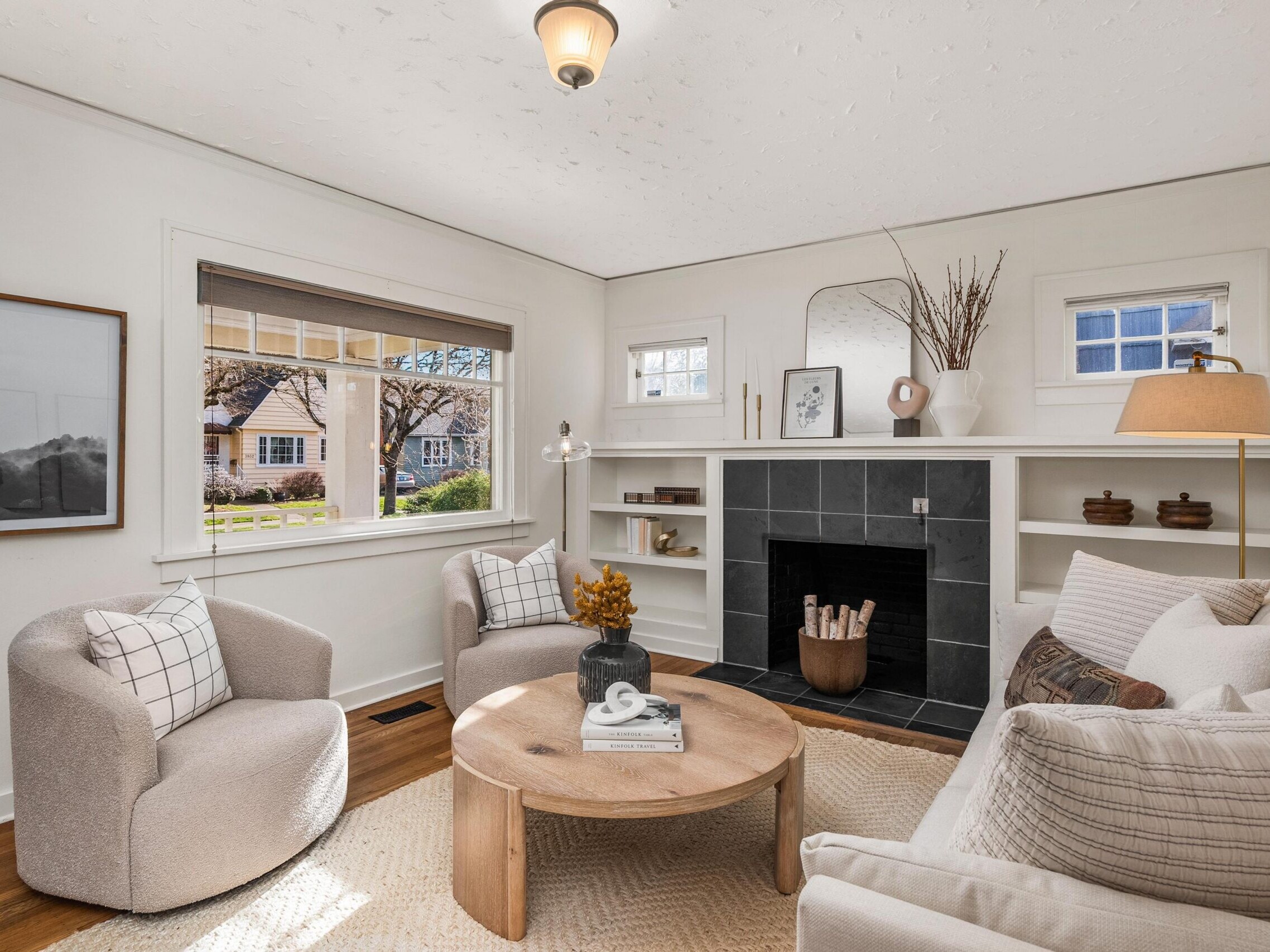 A cozy living room with light walls, two armchairs, a sofa, a round wooden coffee table, and a fireplace with built-in shelves. Large windows let in natural light and decorative items are neatly arranged.