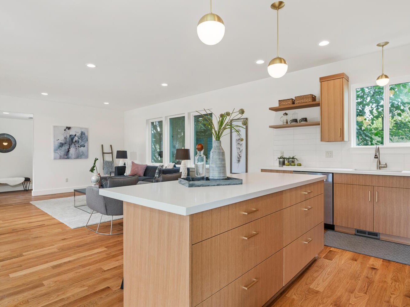 Modern open-concept kitchen and living area with wood floors, light wood cabinets, white countertops, pendant lights, and large windows letting in natural light. A cozy living room is visible in the background.