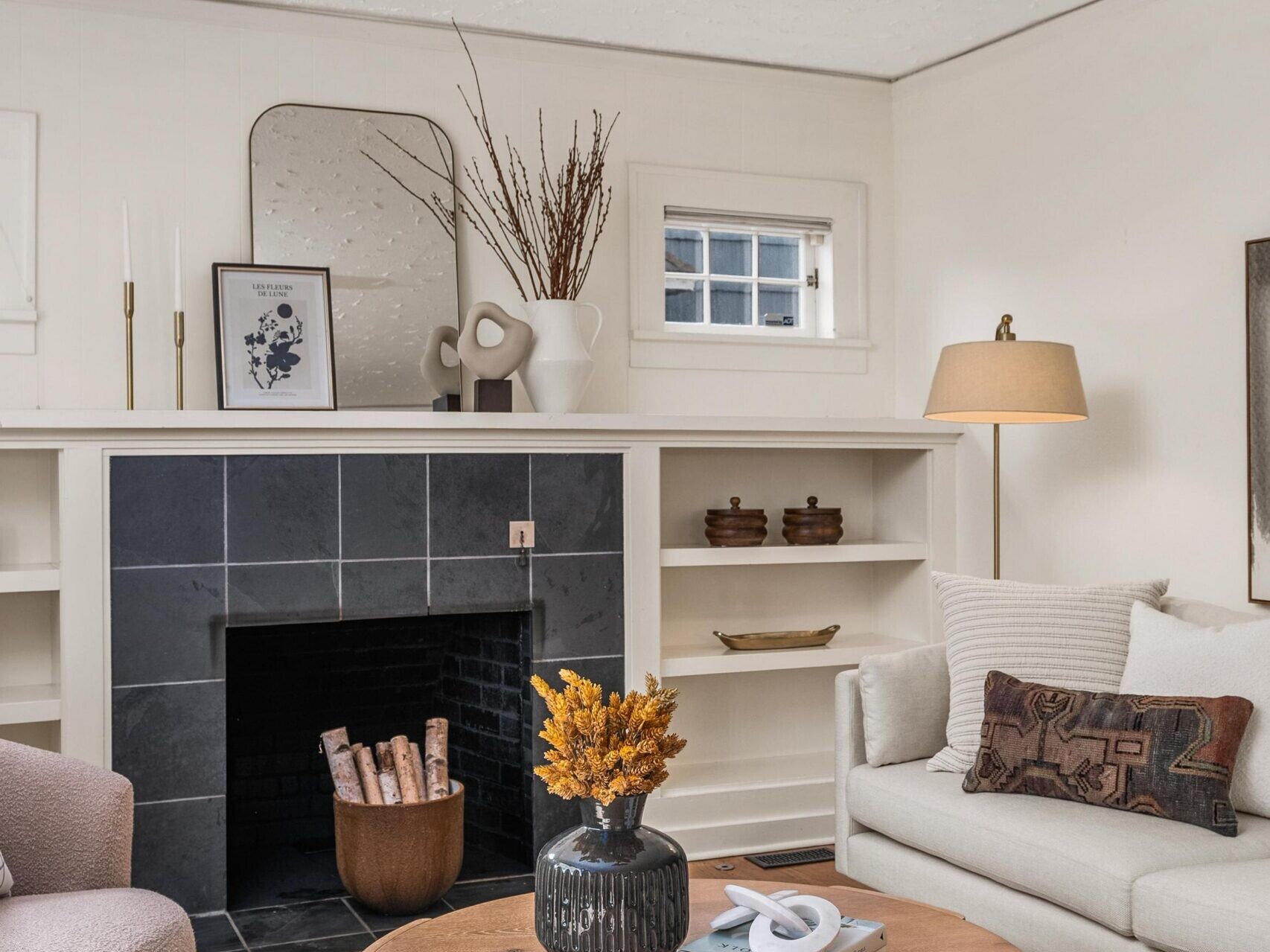A cozy living room with a gray-tiled fireplace, white shelves, a round wooden coffee table with a vase and books, a light sofa with a patterned cushion, light armchair, and neutral decor.