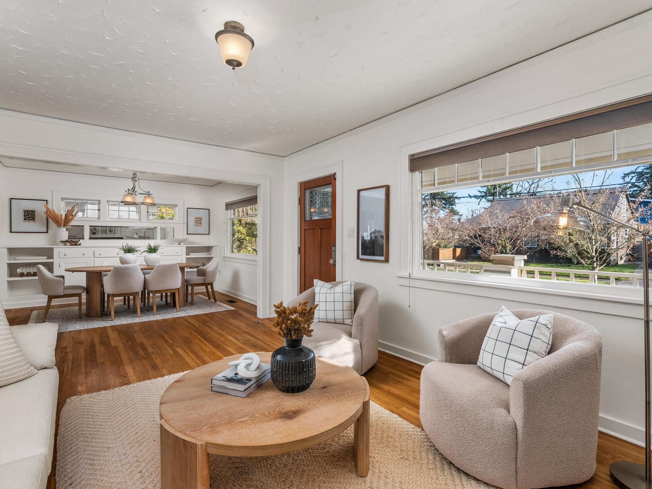 Bright living room with a wooden floor, white walls, and large windows. Cozy seating includes a sofa and two armchairs around a round coffee table. Dining area with six chairs and a light fixture is visible in the background.
