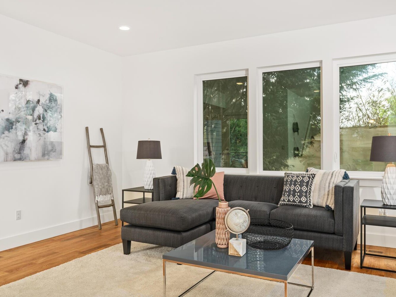 Modern living room with a dark gray sectional sofa, patterned pillows, and a glass coffee table. Two table lamps sit on white side tables. Large windows let in natural light, and a blanket ladder leans against the wall.