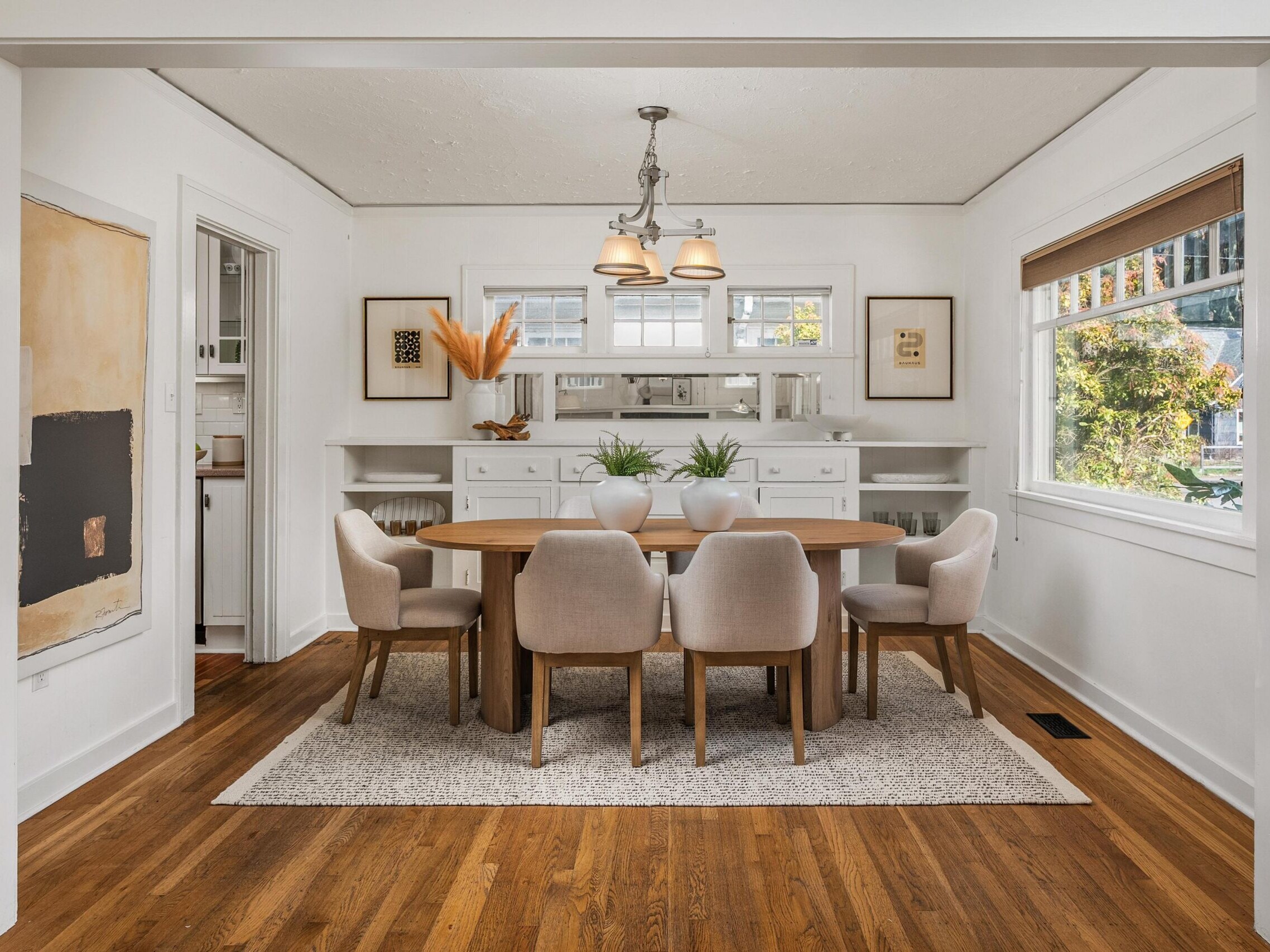 A bright dining room with six beige chairs around a wooden table on a rug, hardwood floors, white built-in cabinets, and large windows letting in natural light. Simple decor with plants and framed art on the walls.