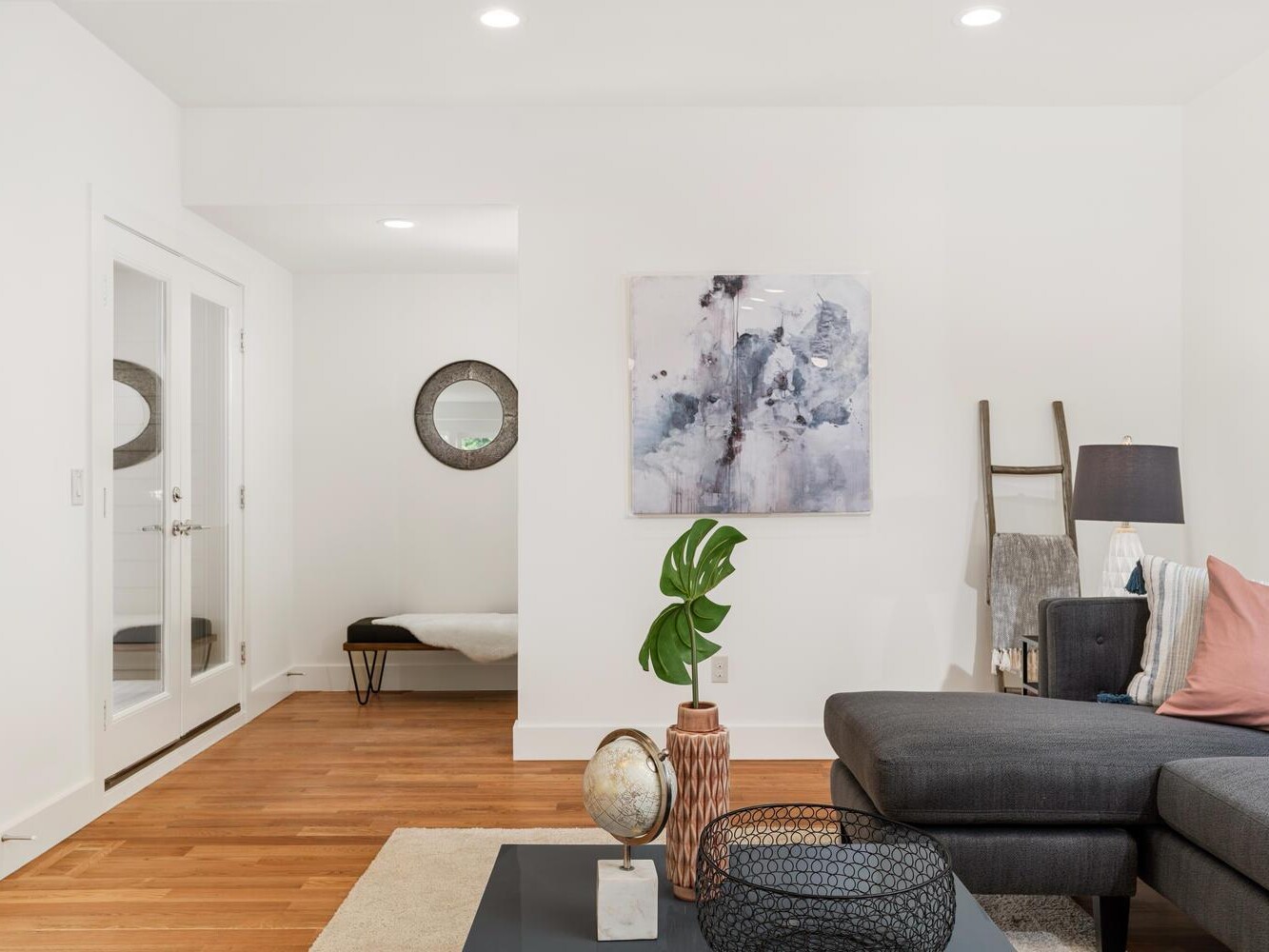 A modern living room with white walls, wooden floors, a gray sectional sofa with pink and gray cushions, abstract wall art, and decorative items on a black coffee table. A ladder shelf and bench sit in the background.