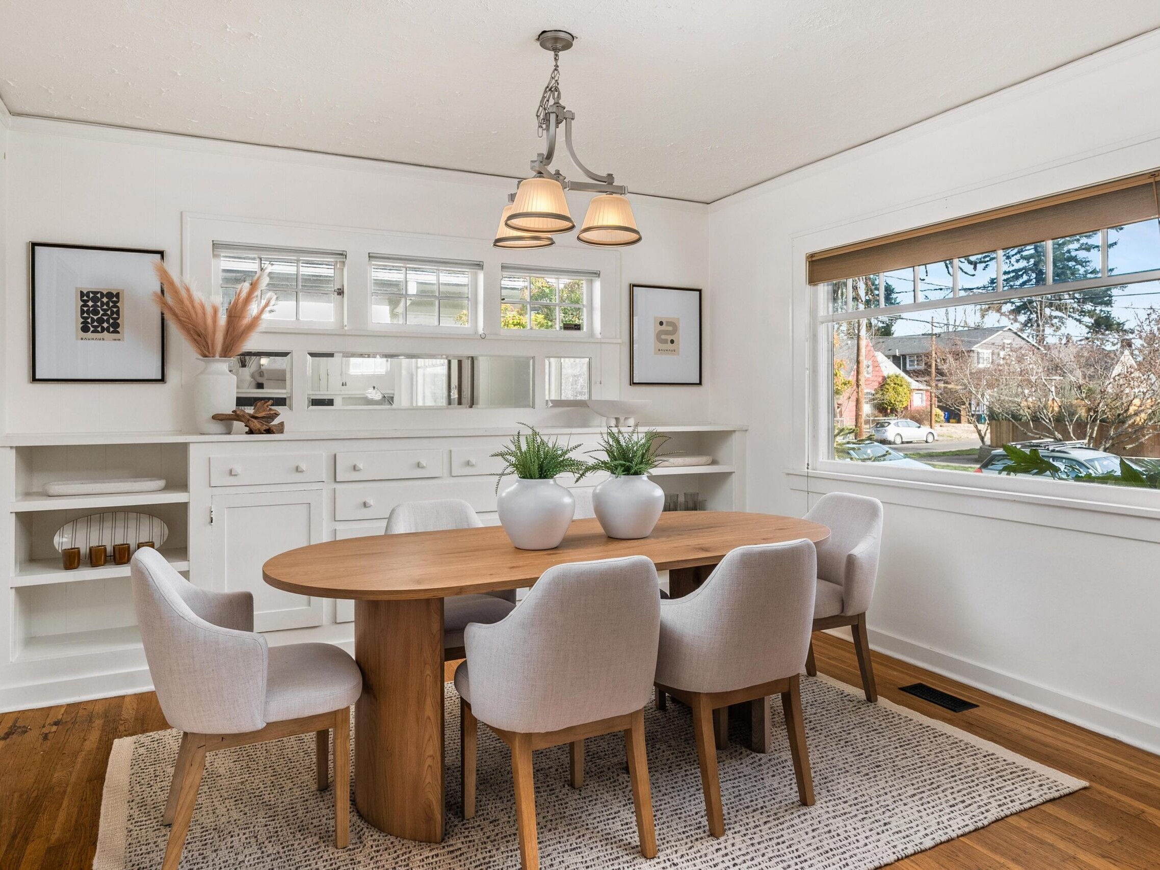 A bright dining room with a wooden table, six light gray chairs, three white vases with green plants, built-in shelves and cabinets, a large window, and a beige rug on a wood floor.
