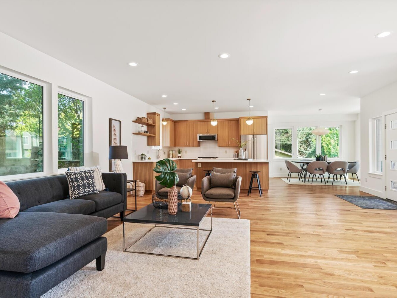 Open-concept living area with wooden floors, a black sectional sofa, modern decor, a kitchen with wood cabinets and island, and a dining table by large windows letting in natural light.