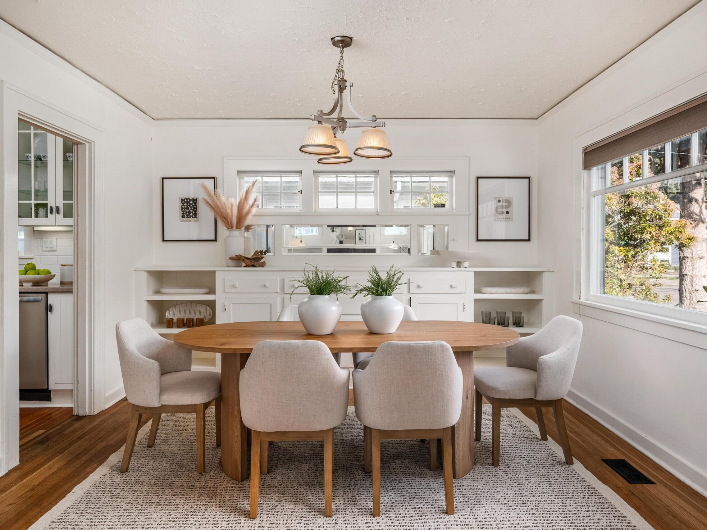 A bright dining room with a wooden table, six light upholstered chairs, two potted plants, white built-in cabinets, framed art on the walls, and large windows letting in natural light.