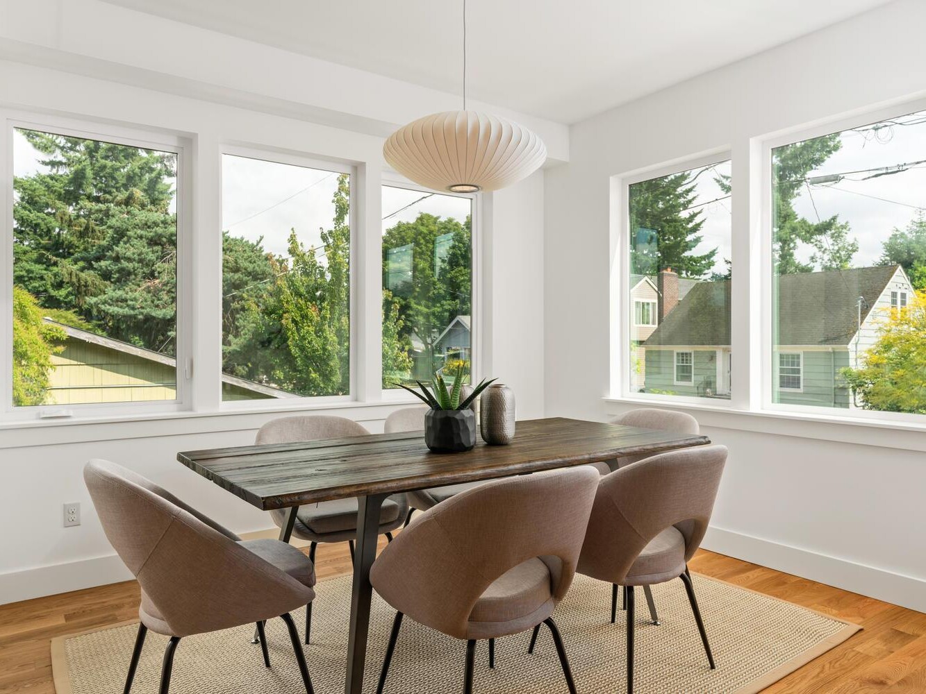 Bright dining room with large windows, a dark wooden table, six beige chairs, a light-colored rug, a modern pendant light, and two vases with a plant on the table. Trees and houses are visible outside the windows.
