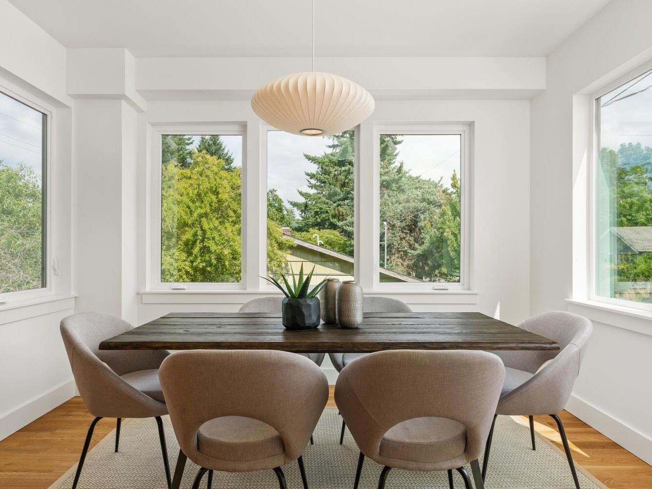 A modern dining room with six beige chairs around a dark wooden table, a white pendant light above, large windows with green trees outside, and vases as a centerpiece on the table.