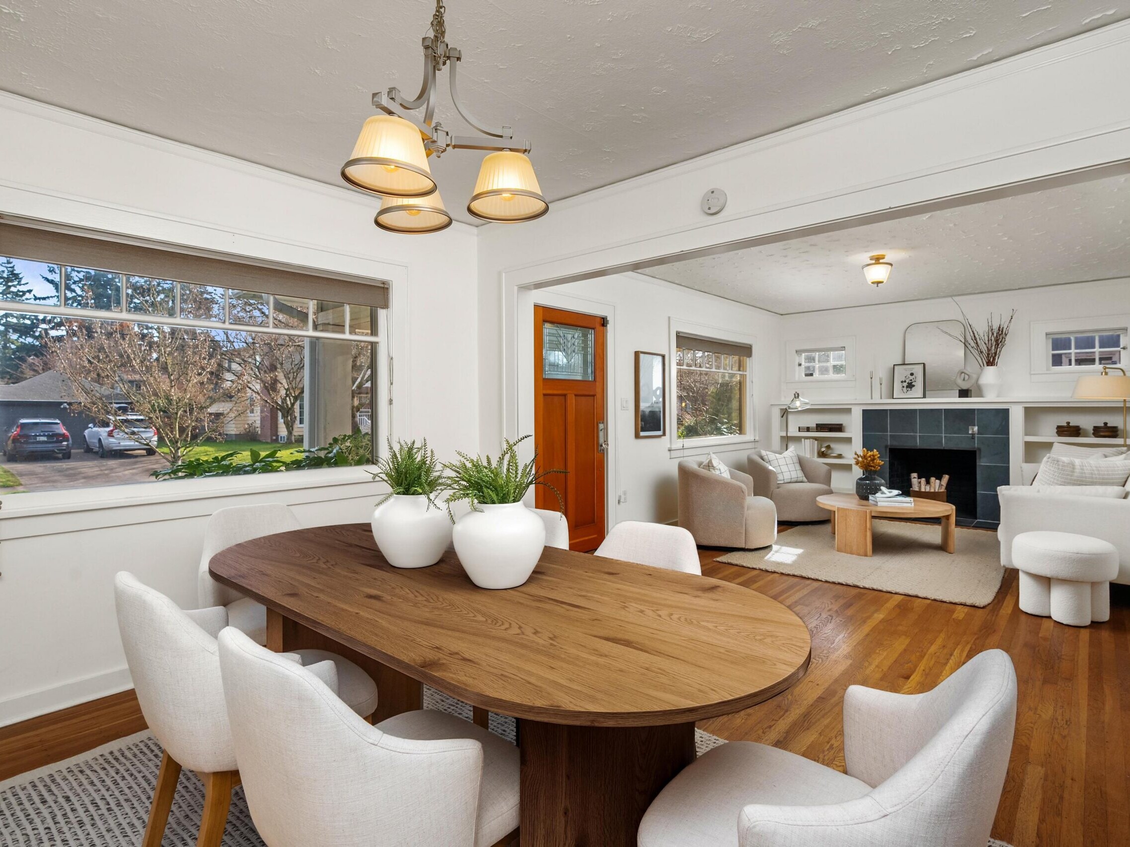 A bright dining area with a wooden table and white chairs is adjacent to a cozy living room with armchairs, a fireplace, and decor. Large windows let in natural light, and a wooden front door is visible.