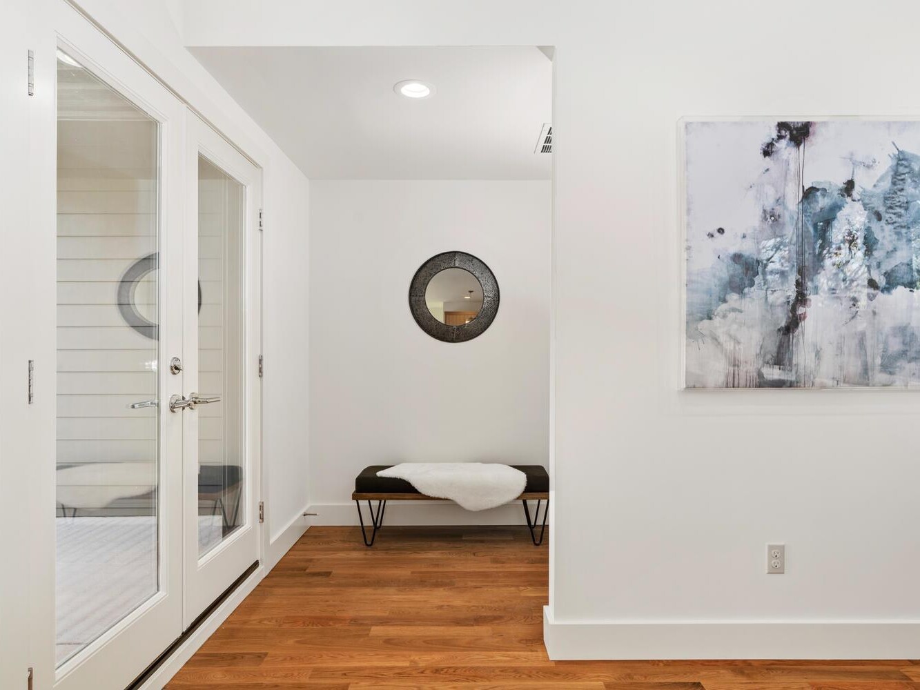 A bright hallway with wooden floors, white walls, French doors on the left, a black bench with a white throw under a round mirror, and an abstract painting hanging on the right wall.