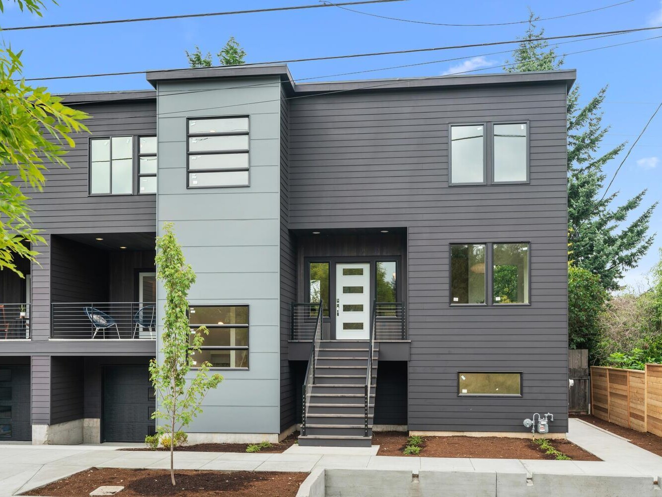 Modern two-story townhouse with dark gray siding, large windows, and a central staircase leading to a white front door. There is a small young tree in front and wooden fencing along the side yard.