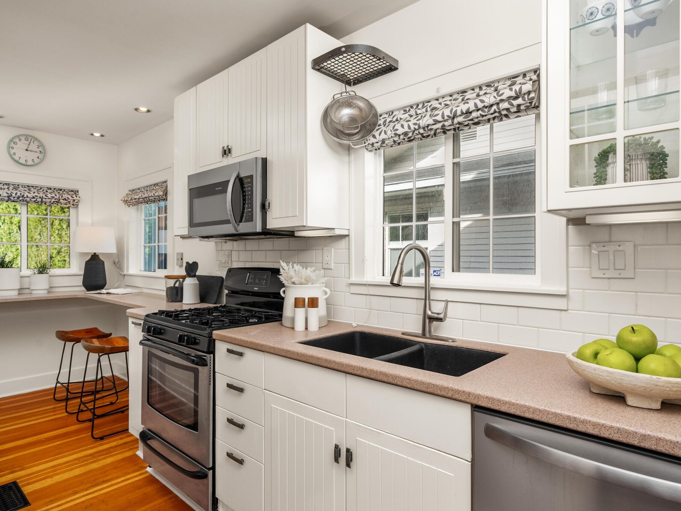 Bright, modern kitchen with white cabinets, stainless steel appliances, a double sink, a bowl of green apples, wood floors, and a breakfast bar with two stools near a window.