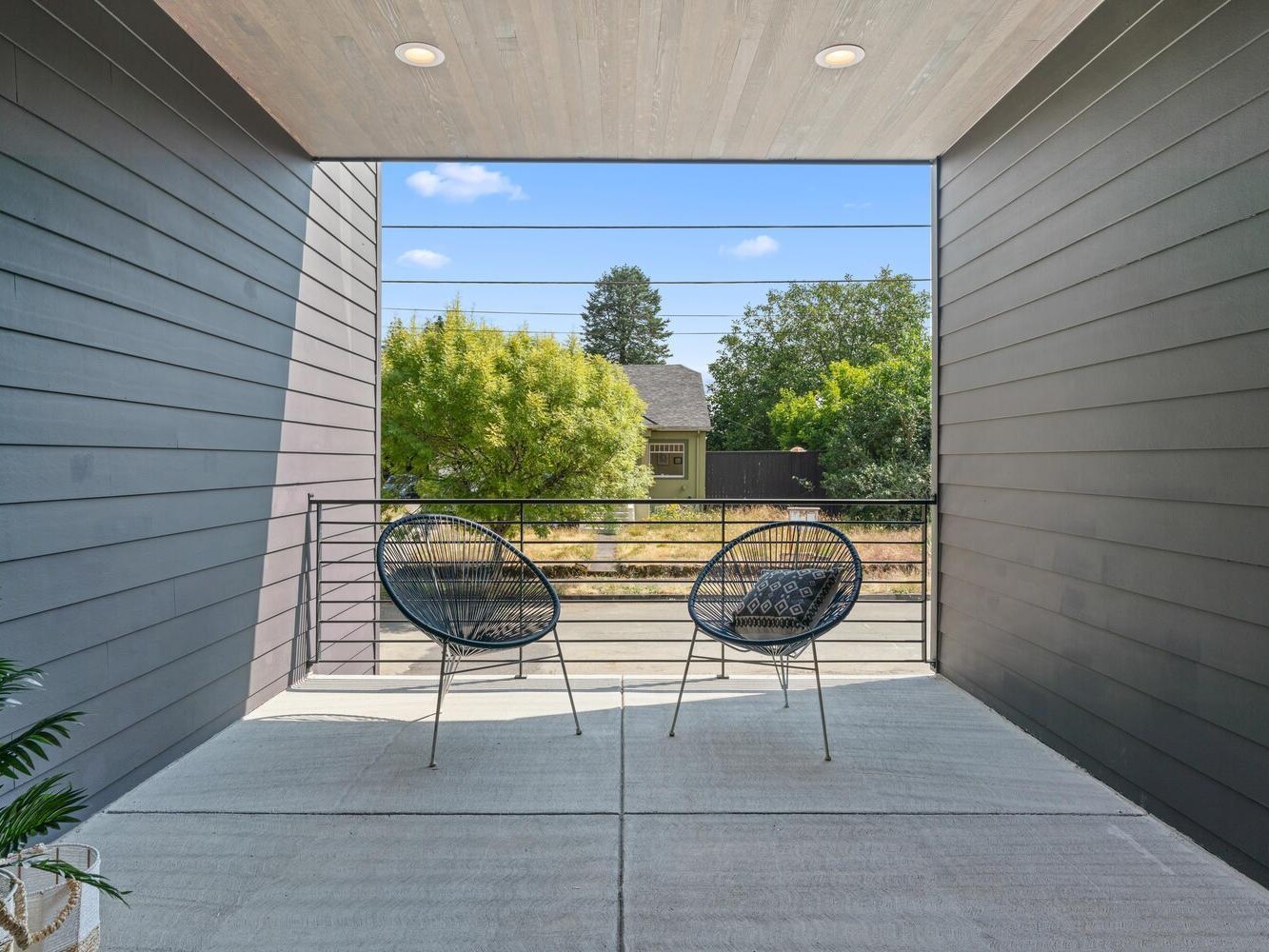 A modern covered balcony with two black wire chairs facing outward, overlooking a residential street with trees and a house in the background on a sunny day.