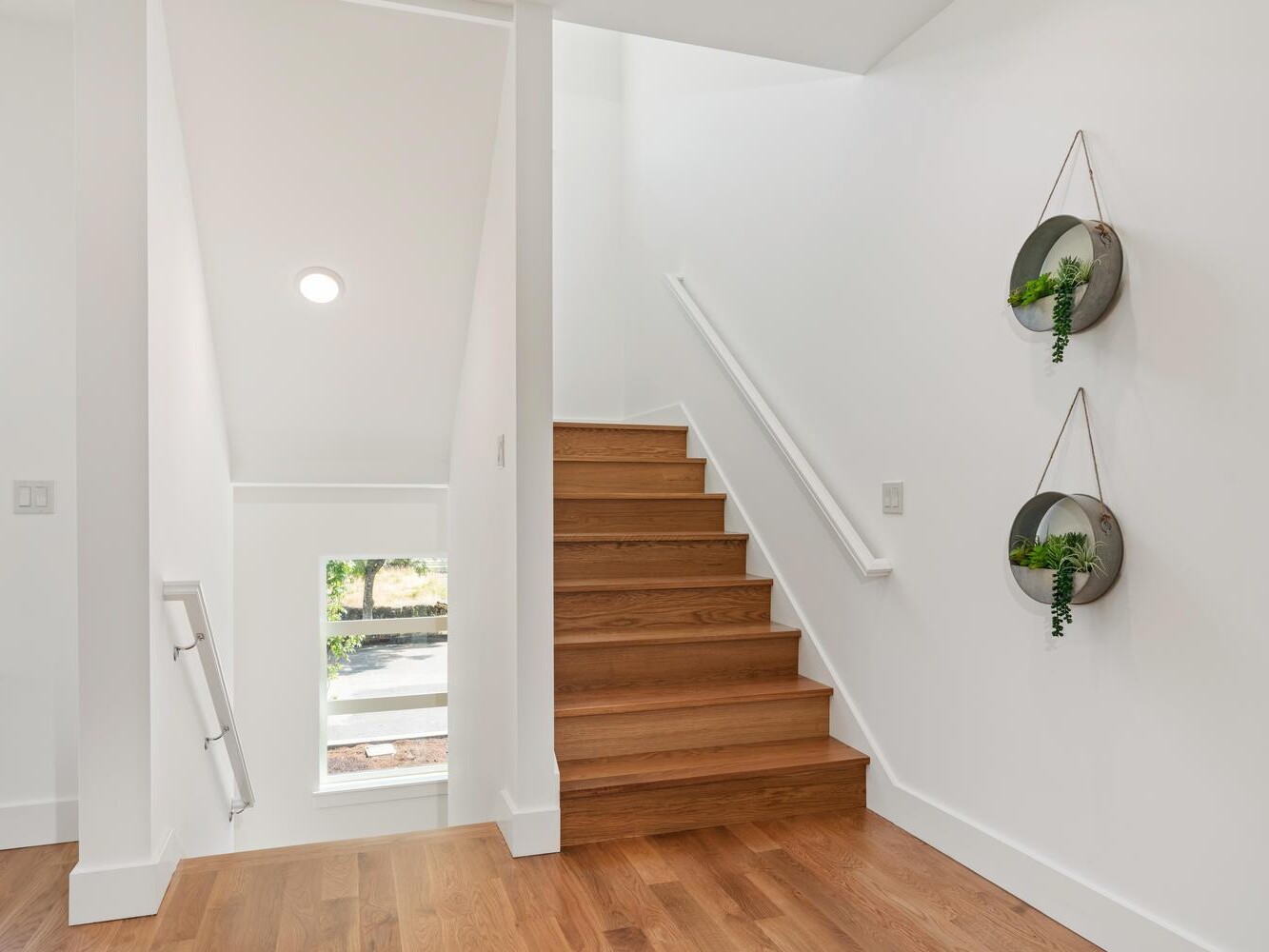 A modern, bright staircase with wooden steps and white walls. Two round wall planters with green plants hang on the right wall. Large windows let in natural light, illuminating the space.