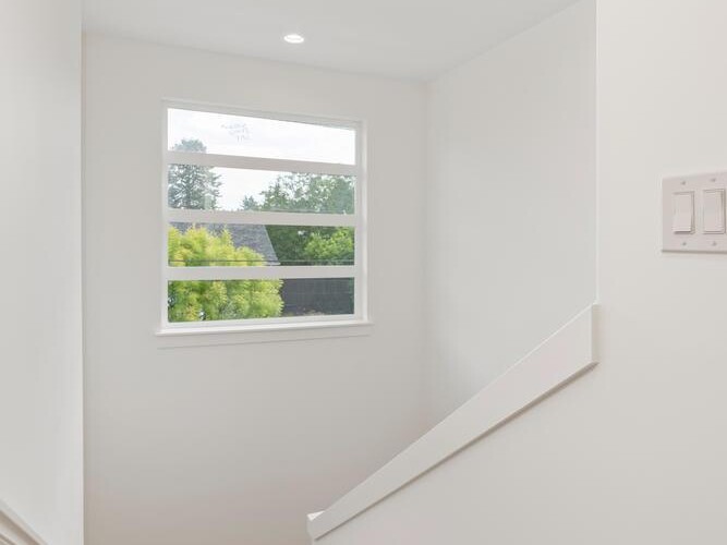 A staircase with wooden steps leads down past a white wall and railing, with a window at the landing letting in natural light and offering a view of trees and a building outside.