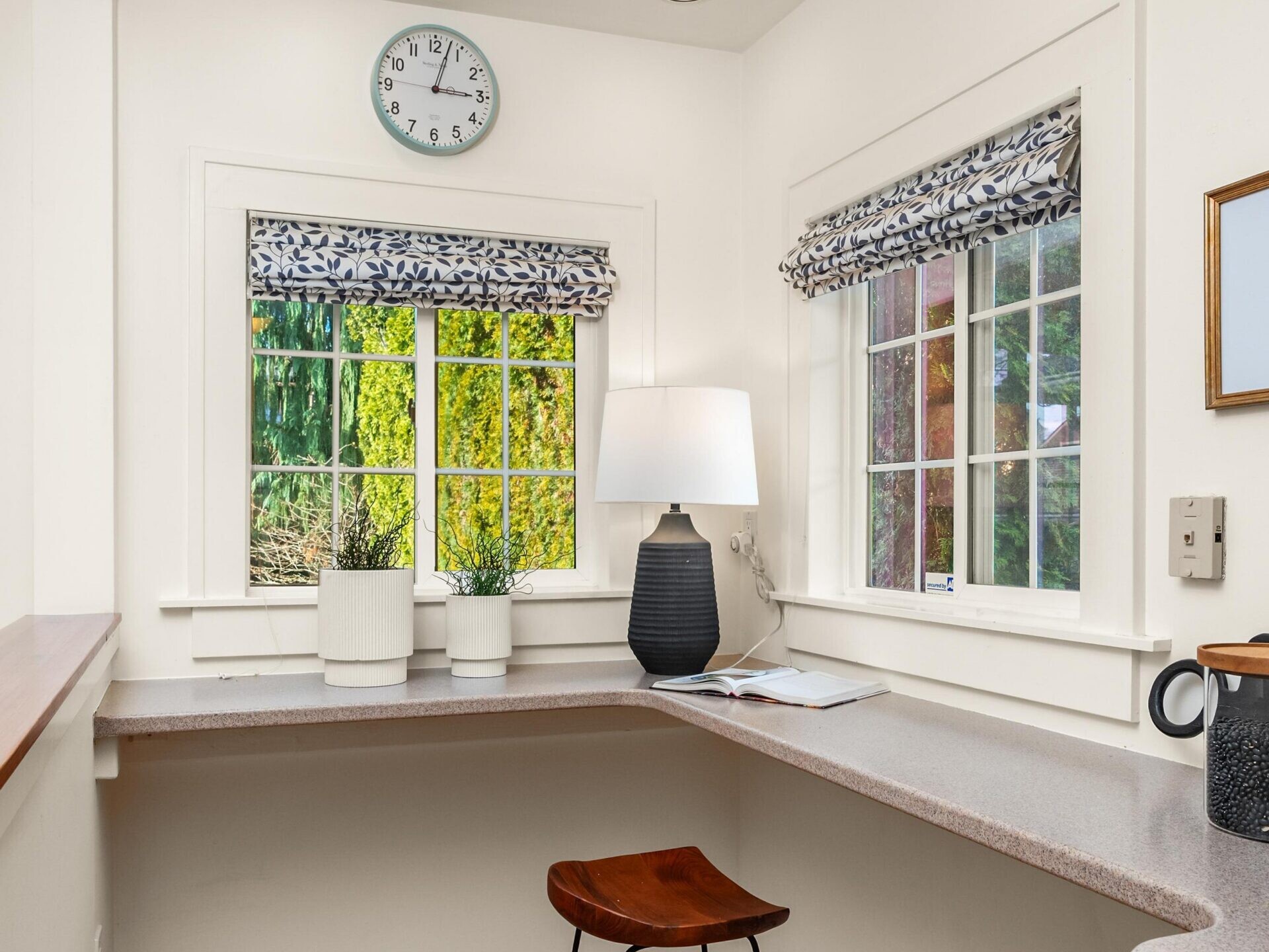 A small kitchen corner with a built-in counter, two wooden stools, a table lamp, potted plants, a magazine, and two windows with patterned blinds showing green trees outside. A wall clock is above the windows.