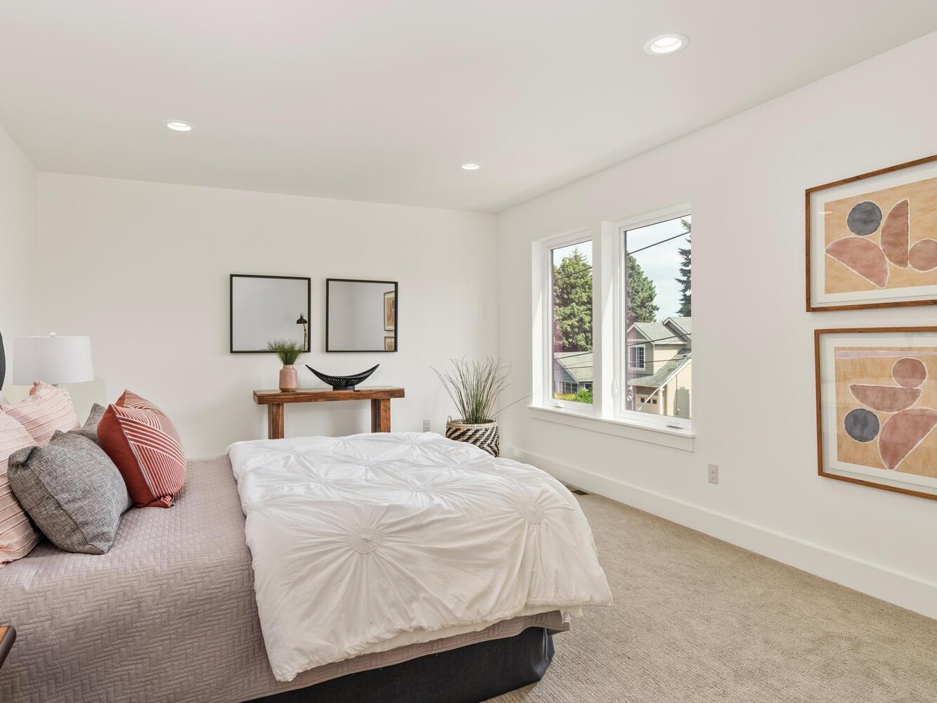 Modern bedroom with a neatly made bed, decorative pillows, light bedding, wall art, a wooden console table with decor, two windows letting in natural light, and neutral-colored walls and carpet.