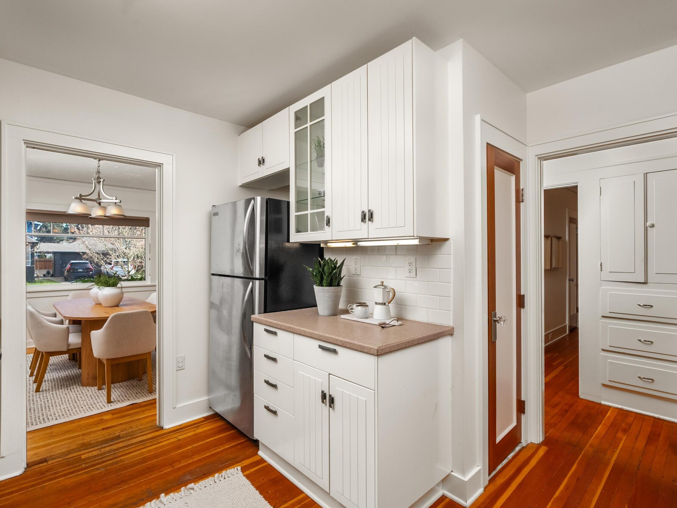Bright kitchen with white cabinets, stainless steel refrigerator, brown countertops, and hardwood floors. A plant sits on the counter. Dining room with table and chairs visible through a doorway.