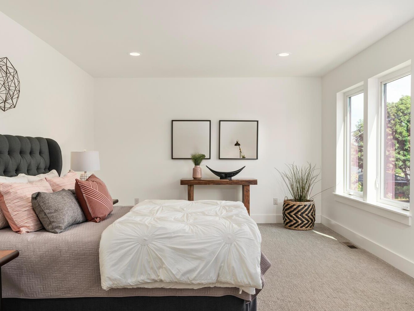 A modern, bright bedroom with a tufted dark gray headboard, white and gray bedding with accent pillows, a wooden desk with decor items, wall art, and large windows letting in natural light.