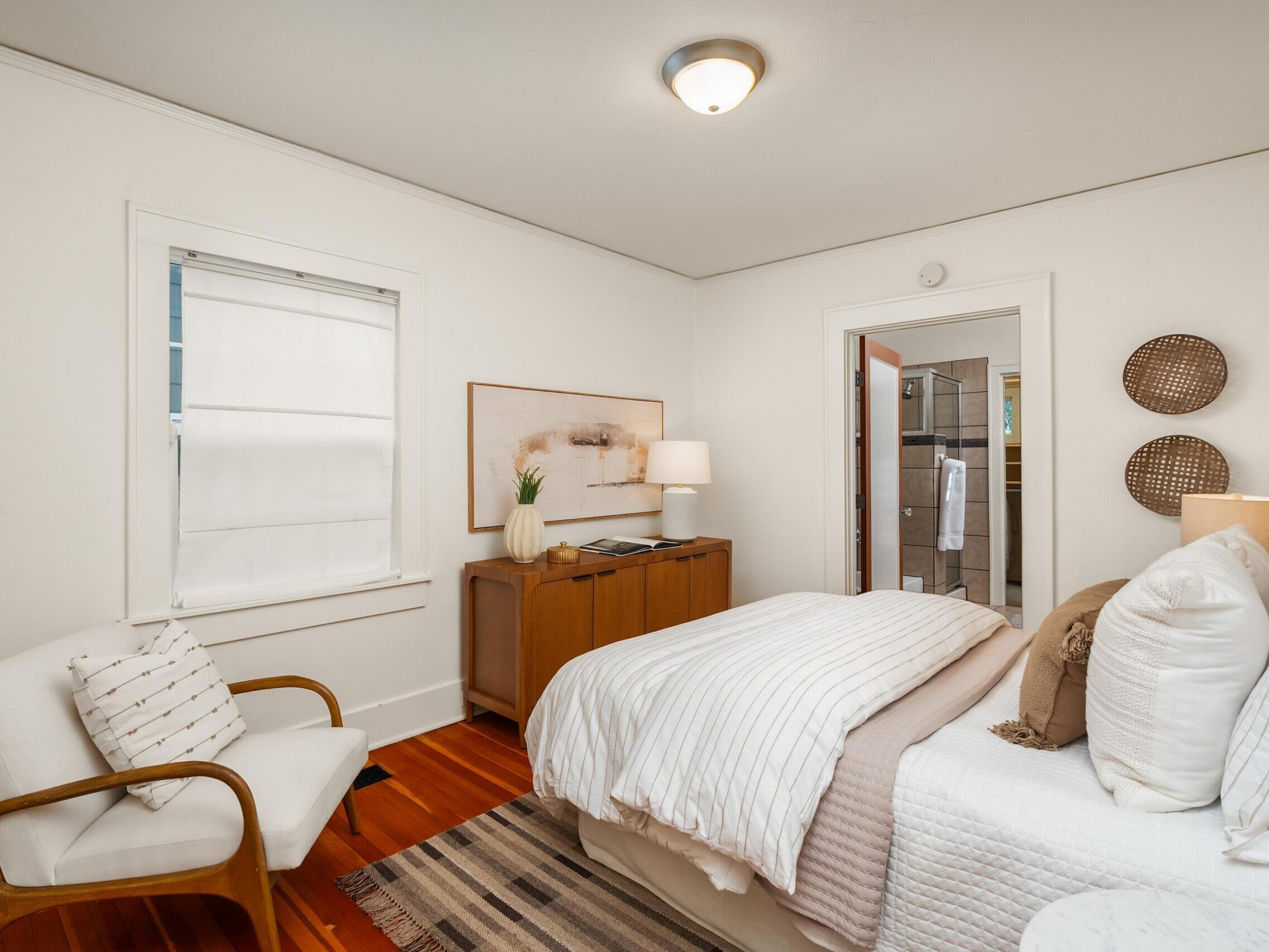 A cozy bedroom with a neatly made bed, white walls, wooden floors, a small wooden dresser with a lamp, a white cushioned chair, and a window with a white shade. The door reveals a bathroom beyond.