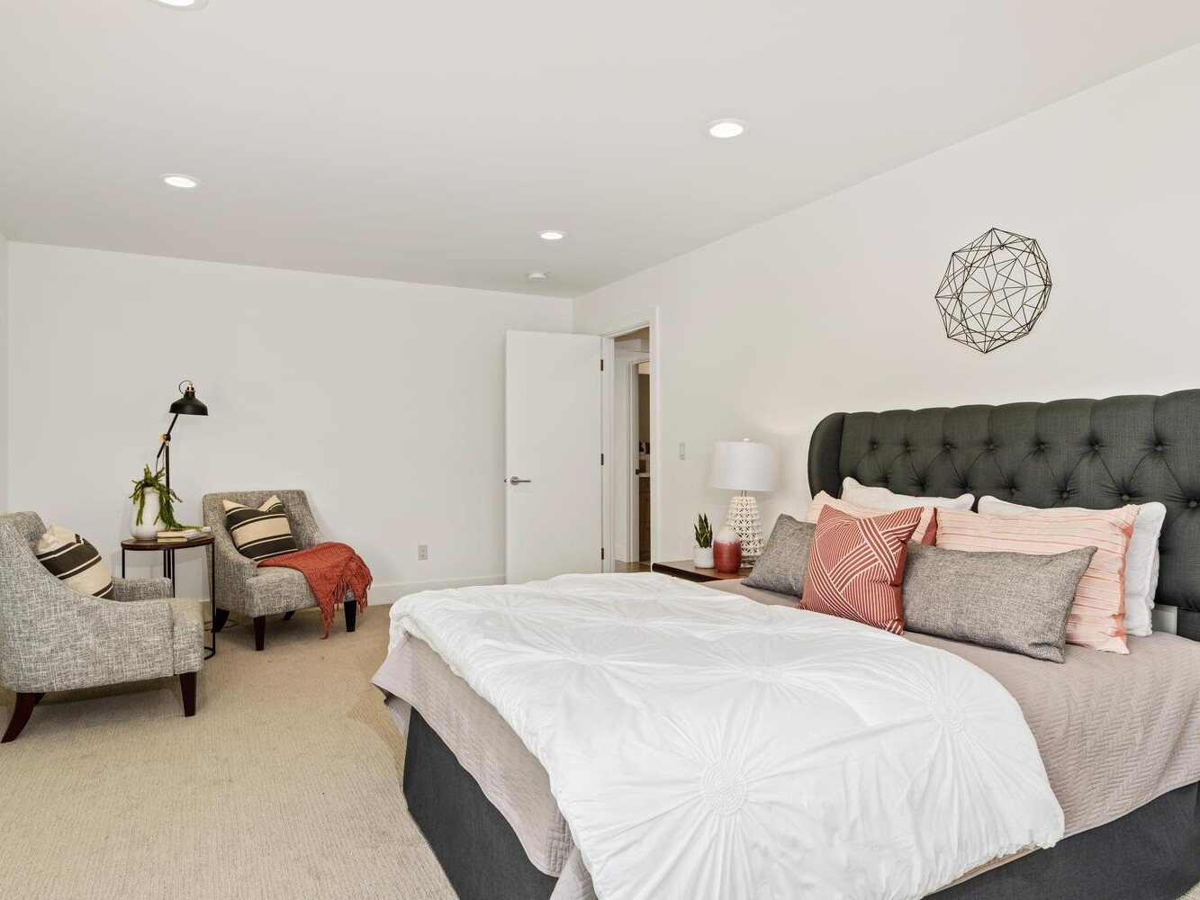 Modern bedroom with a large upholstered bed, white bedding, and multiple pillows. Two armchairs with cushions and throw blankets sit in the corner near a floor lamp and small plant. Walls and décor are minimal and light-toned.