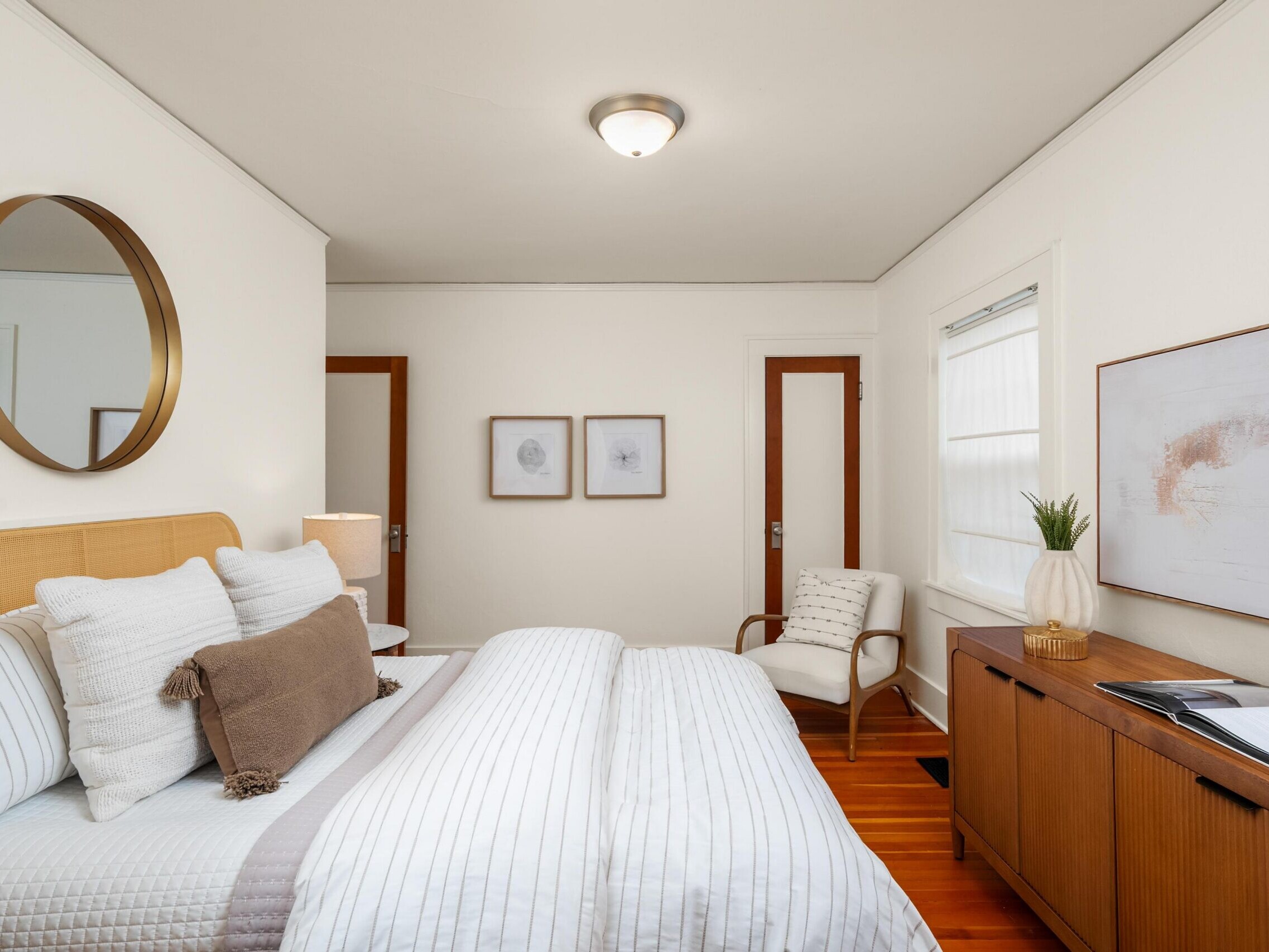 A cozy, modern bedroom with a neatly made bed, striped bedding, two framed artworks on a white wall, a wooden sideboard with decor, an armchair, and natural light from a window with blinds.