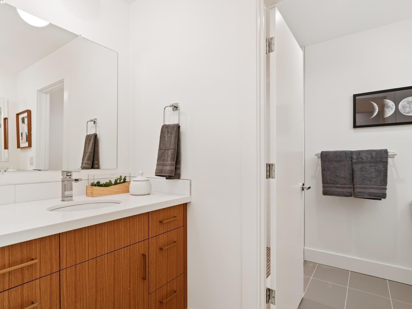 A modern bathroom with a wooden vanity, white countertop, and rectangular mirror. In the adjacent room, a toilet and towel rack with dark towels are visible, along with a framed moon phase print on the wall.