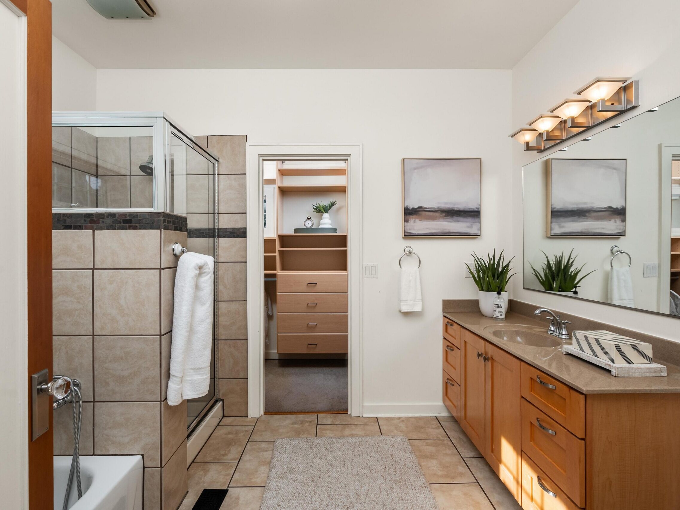 Modern bathroom with a glass-enclosed shower, double sink vanity, large mirror, tile floor, neutral decor, and an open doorway leading to a walk-in closet with built-in shelving and drawers.