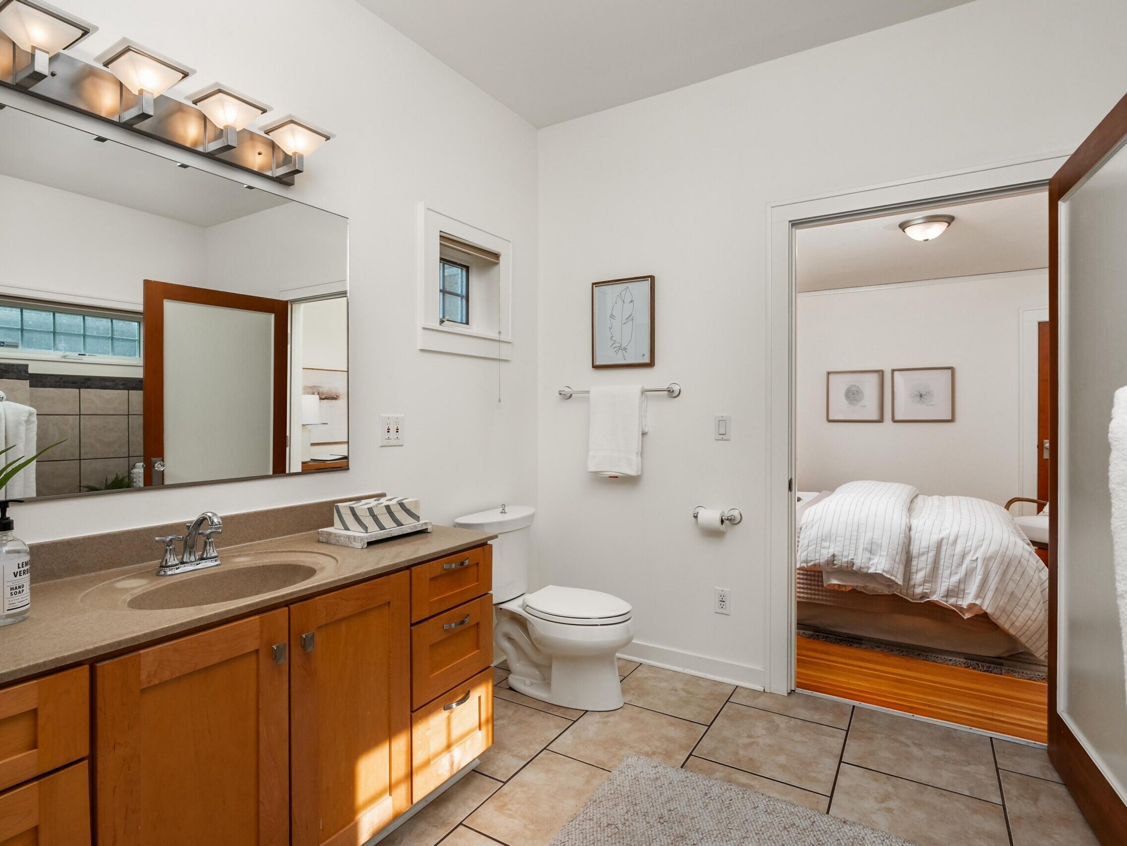 A modern bathroom with a wooden vanity, large mirror, beige countertop, and tile floor. A toilet sits beside a towel rack, and an open door reveals a neatly made bed in an adjoining bedroom.