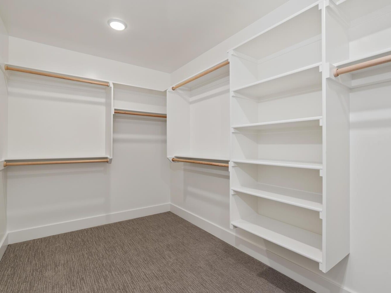 Spacious, empty walk-in closet with white shelves and wooden hanging rods on both sides. The walls and ceiling are white, and the floor is carpeted in a neutral color. Ceiling light fixture is visible.