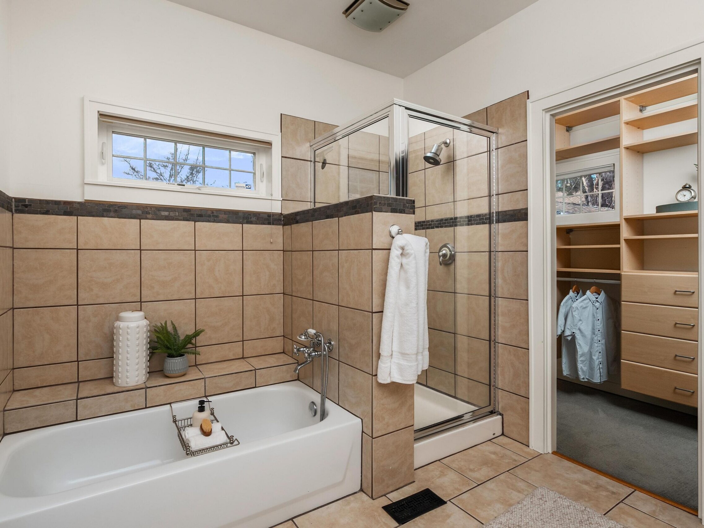 Modern bathroom with beige tile, a bathtub with decorative items, a glass-enclosed shower, towel on the door, and an adjoining walk-in closet with hanging clothes and built-in shelves.