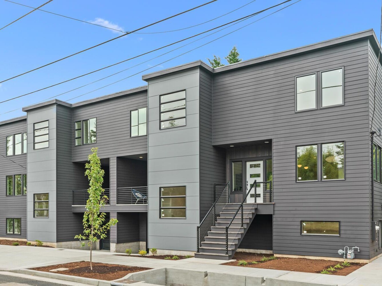 Modern two-story apartment building with dark gray horizontal siding, large windows, a central staircase leading to the entrance, and young landscaping in front, set against a bright blue sky.