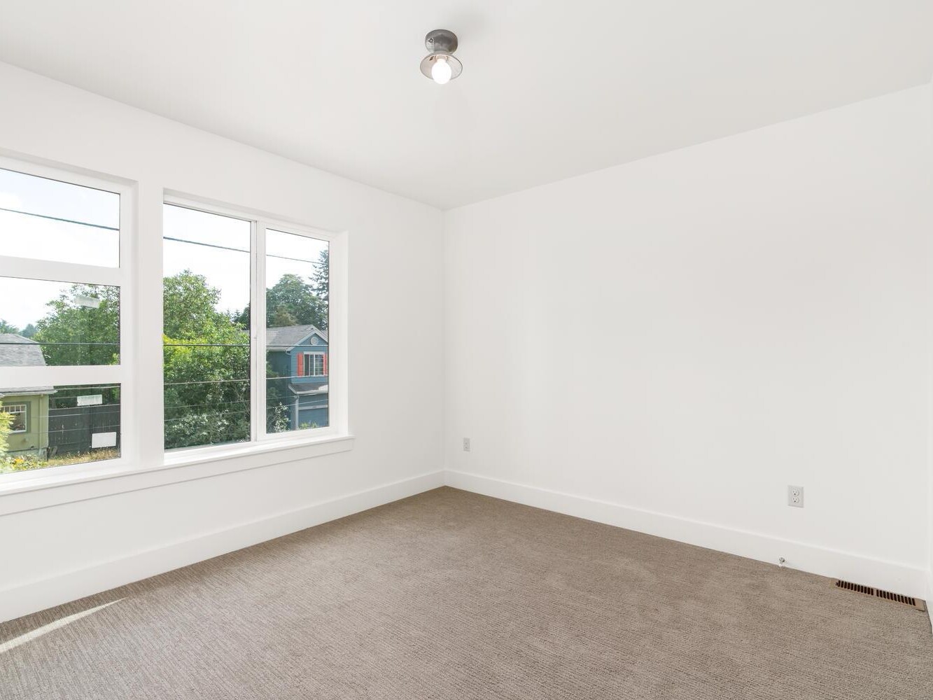 A bright, empty room with white walls, a beige carpet, a ceiling light fixture, and large windows letting in natural light with a view of trees and neighboring houses.