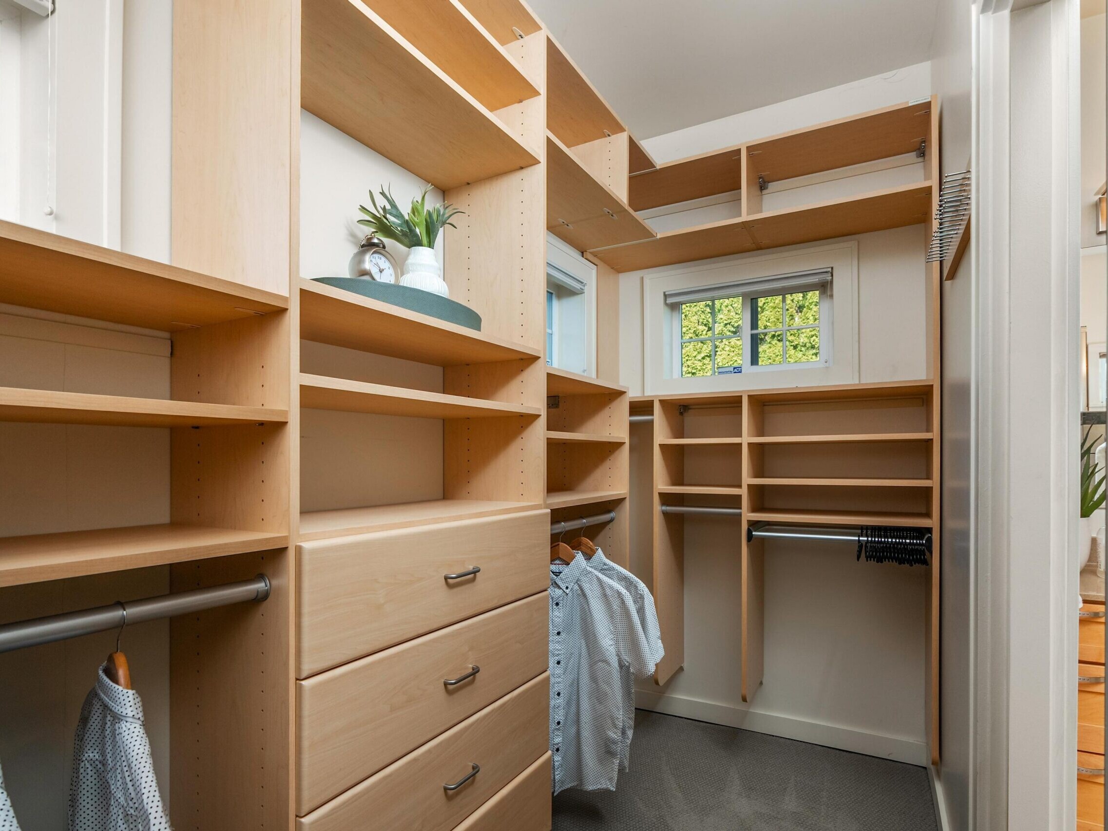 A walk-in closet with light wood shelves, drawers, hanging rods with shirts, and a small window letting in natural light. Some decorative items and plants are on the shelves.