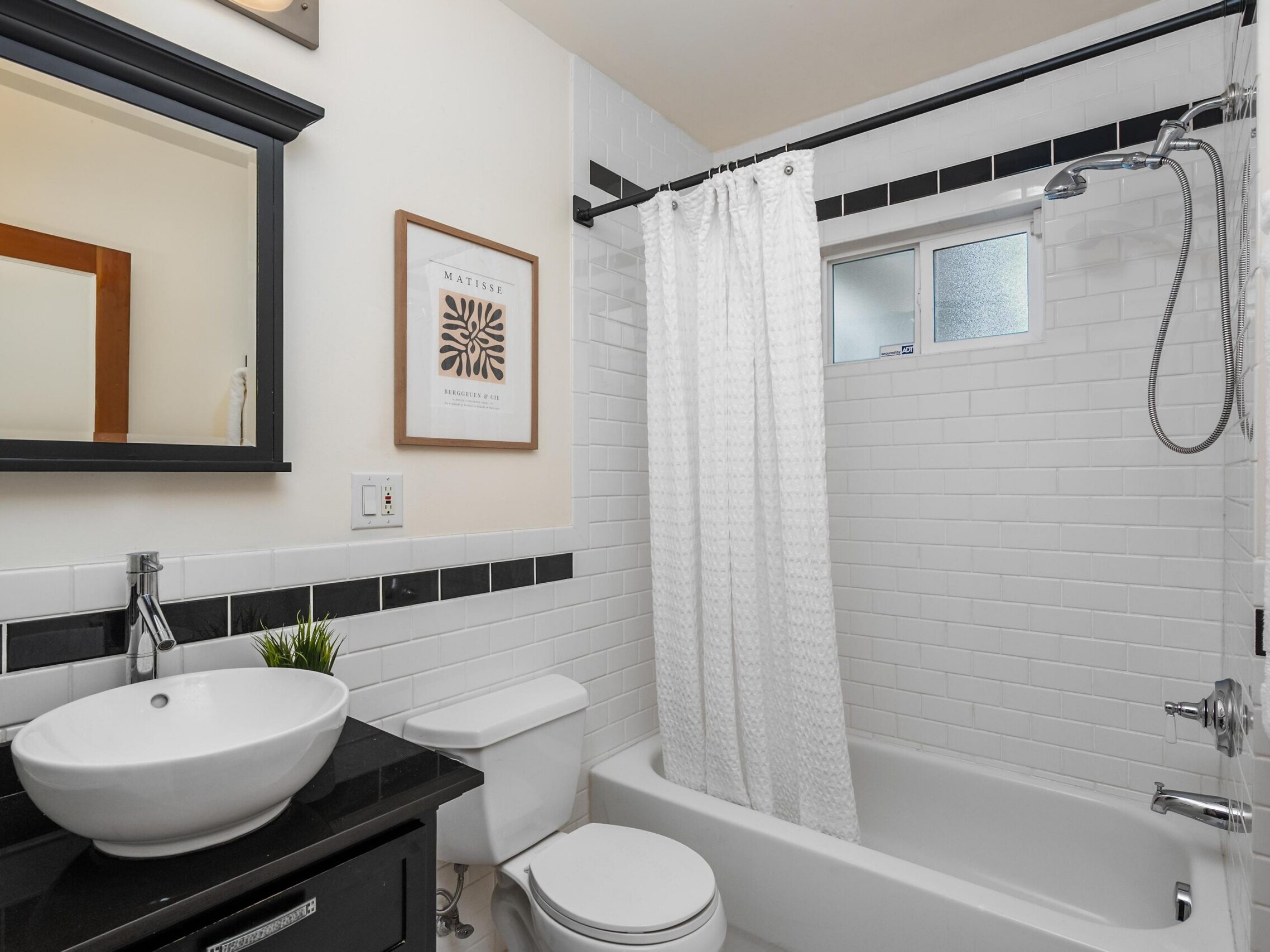A modern bathroom with white subway tile walls, a white bathtub with a shower and curtain, a round sink on a black counter, a toilet, a framed print on the wall, and towels neatly hung.